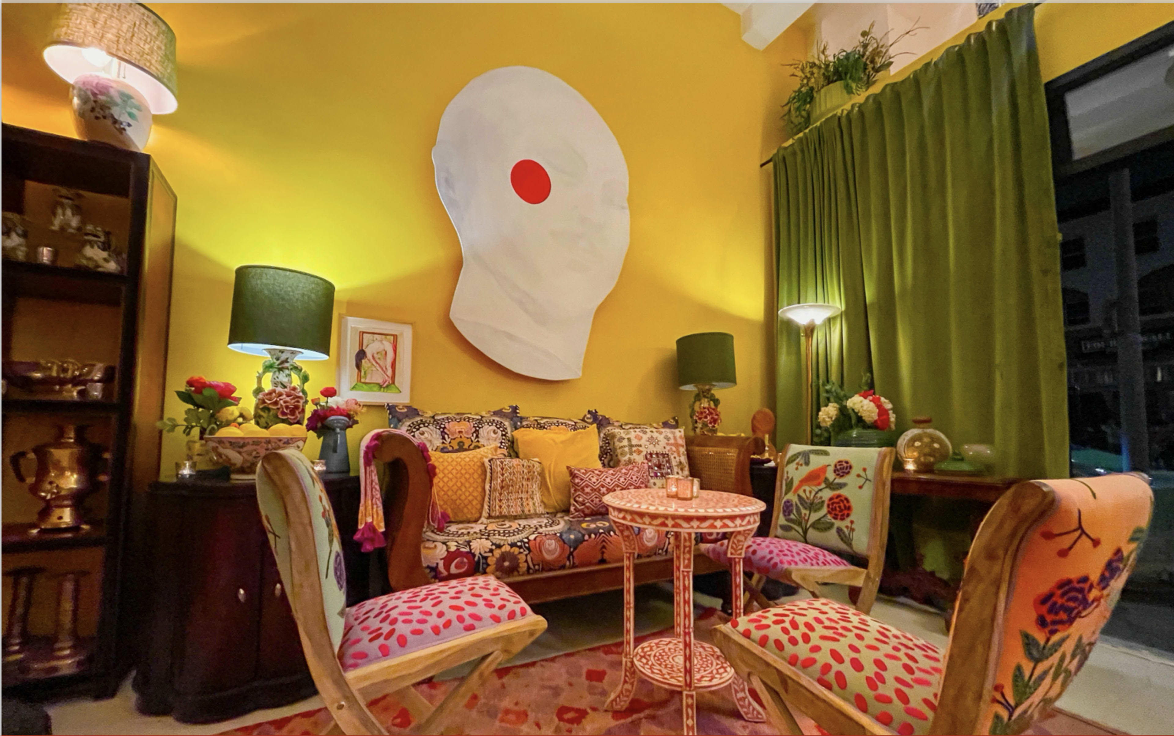 The image shows a colorful interior setting with a yellow wall, a large white art piece resembling a head, and a cozy seating area featuring patterned cushions and a small round table surrounded by floral chairs.