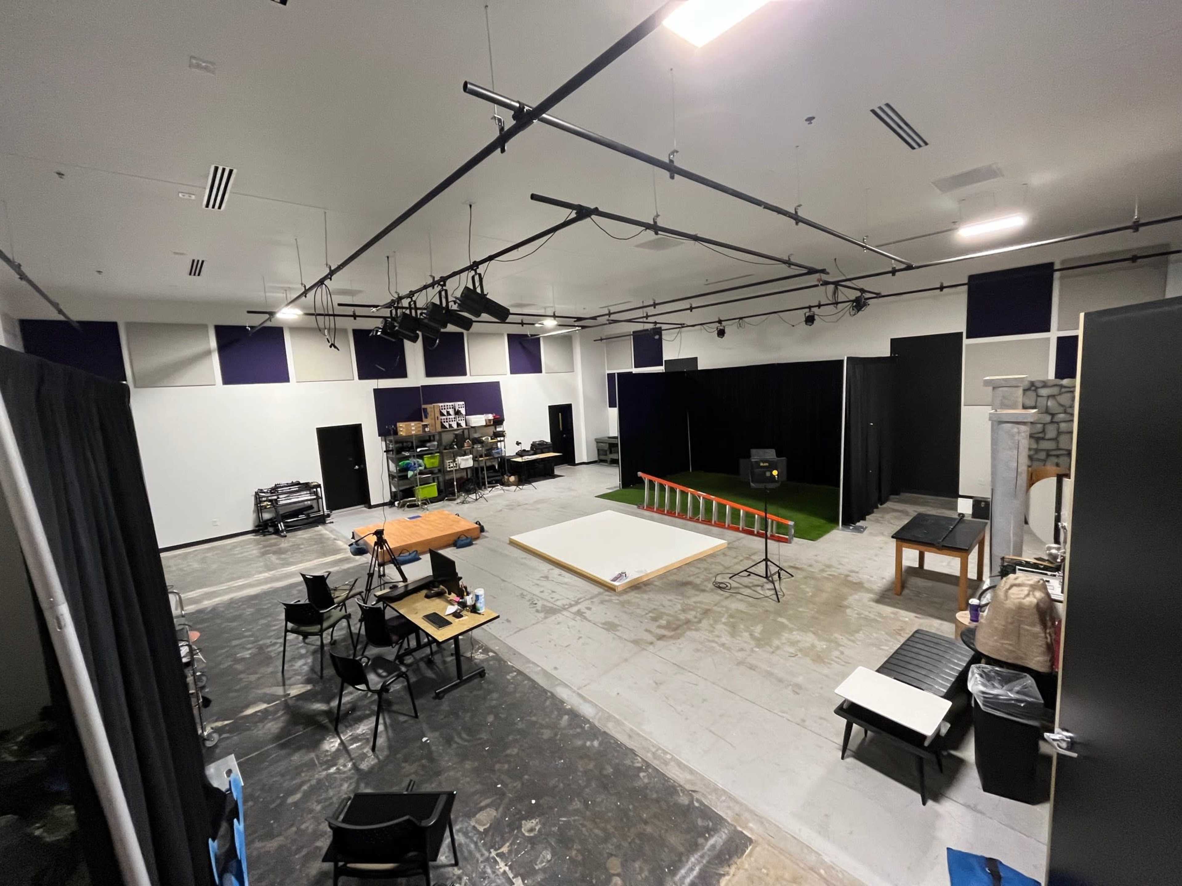 A spacious studio with various equipment, props, and staging areas, featuring tables, chairs, and a small stage with a black curtain backdrop.
