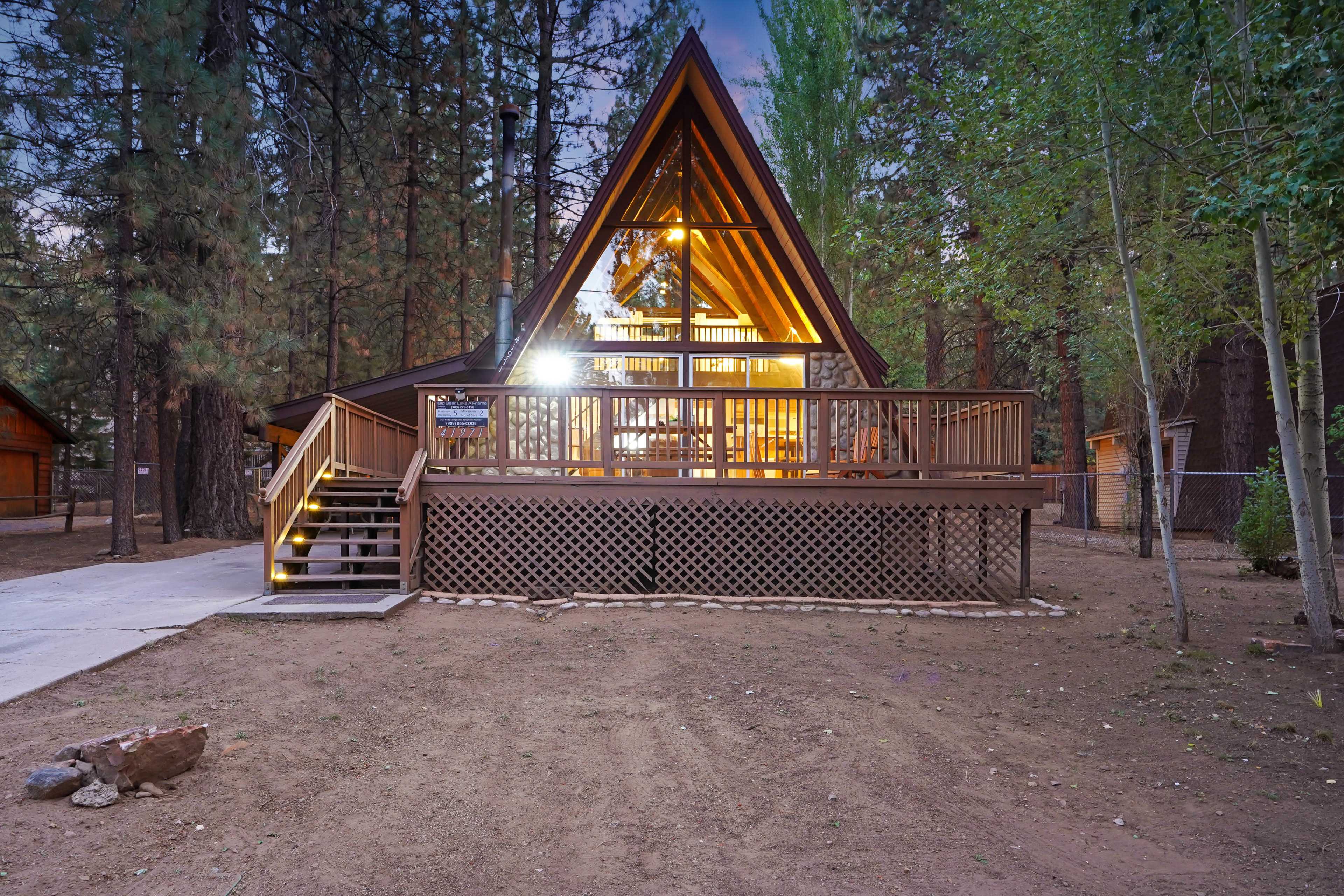 A wooden A-frame cabin with large windows and a deck is surrounded by pine trees in a forested area.