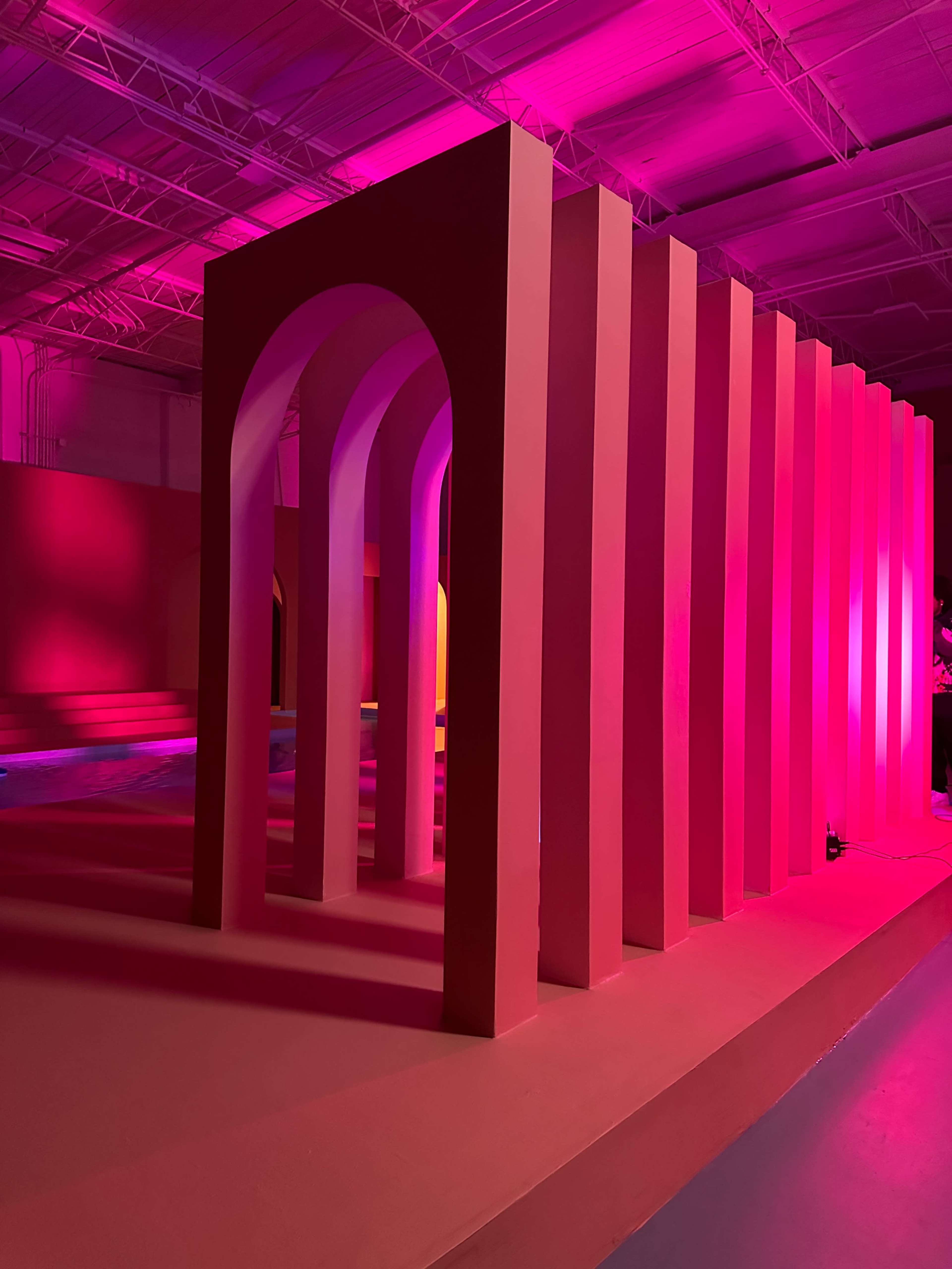 A series of large, colorful arches and vertical structures illuminated by pink lighting in an industrial space.