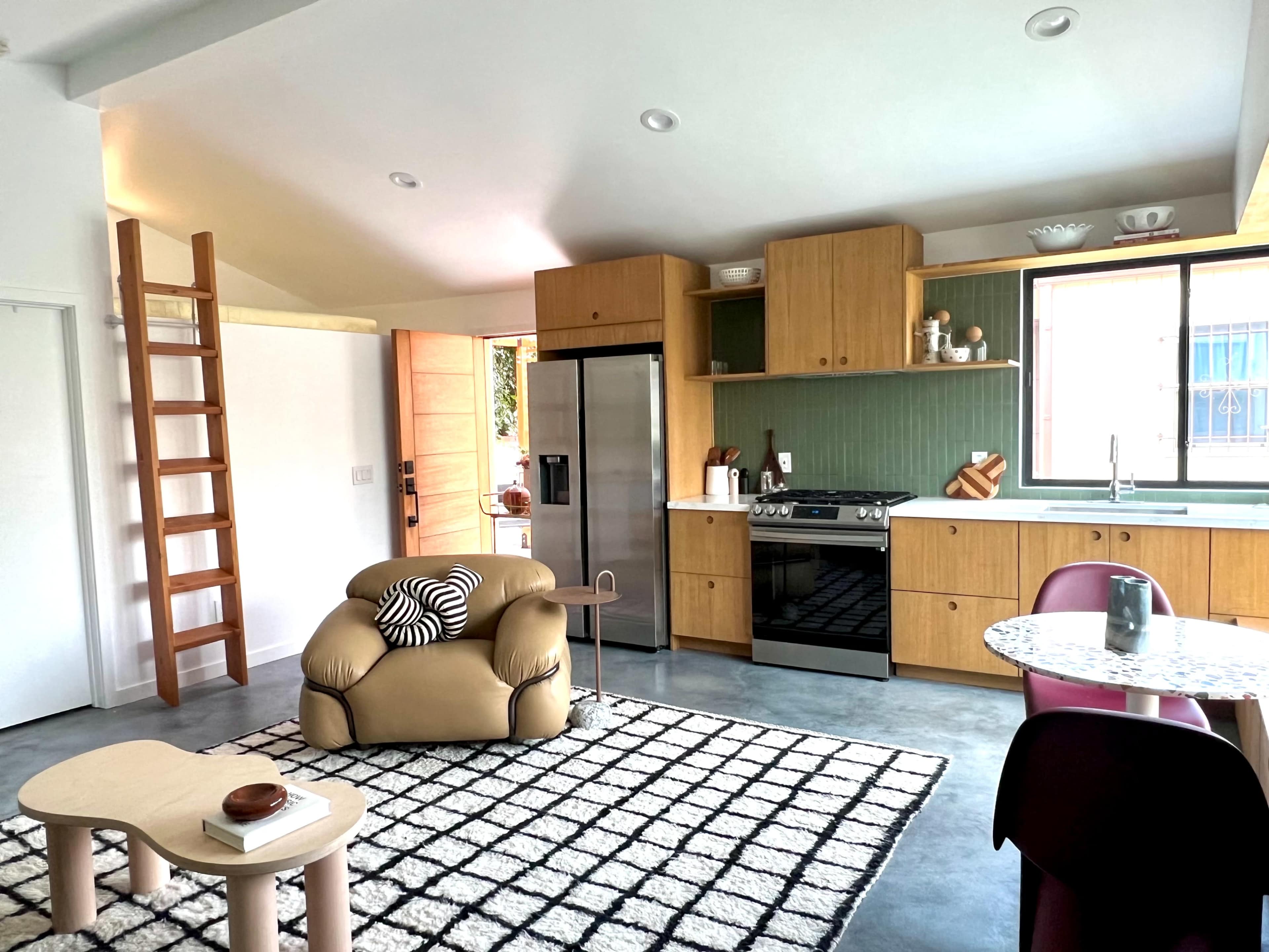 A modern kitchen and living area features wooden cabinetry, a stainless steel refrigerator, and a patterned rug with a cozy chair and dining table.
