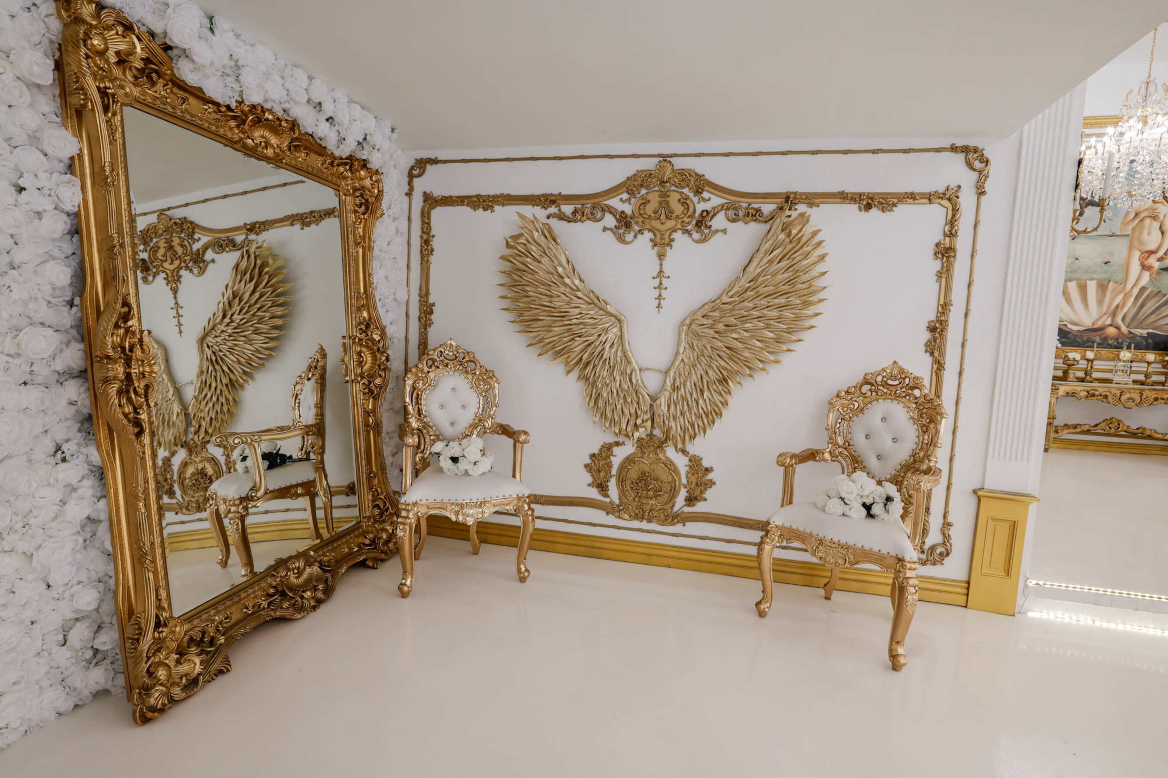 A lavishly decorated room featuring a large ornate mirror, two elegant chairs, and a prominent wall feature with golden angel wings.