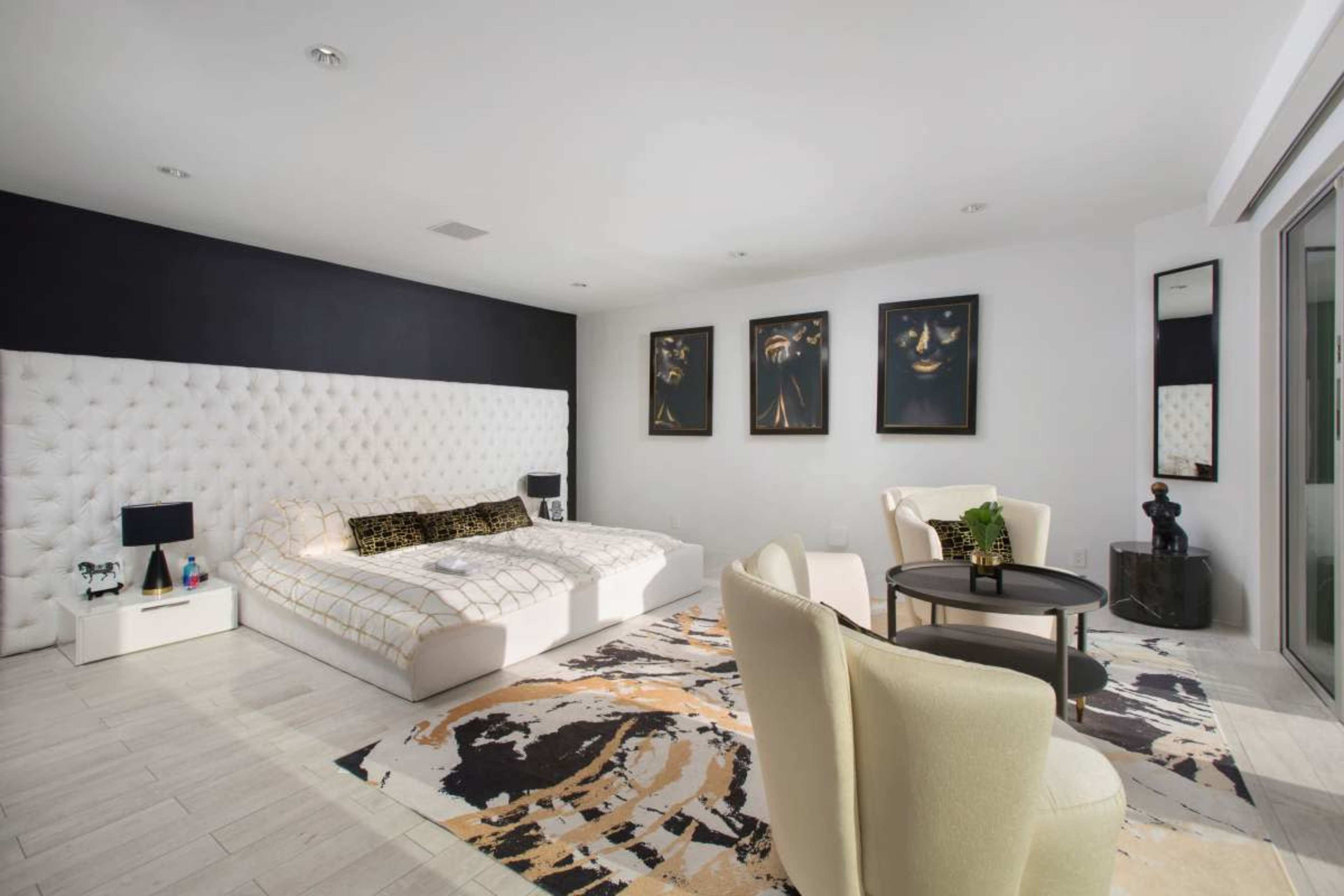 The image shows a modern bedroom featuring a large tufted bed against a black accent wall, complemented by two stylish chairs and a round table on a patterned rug.