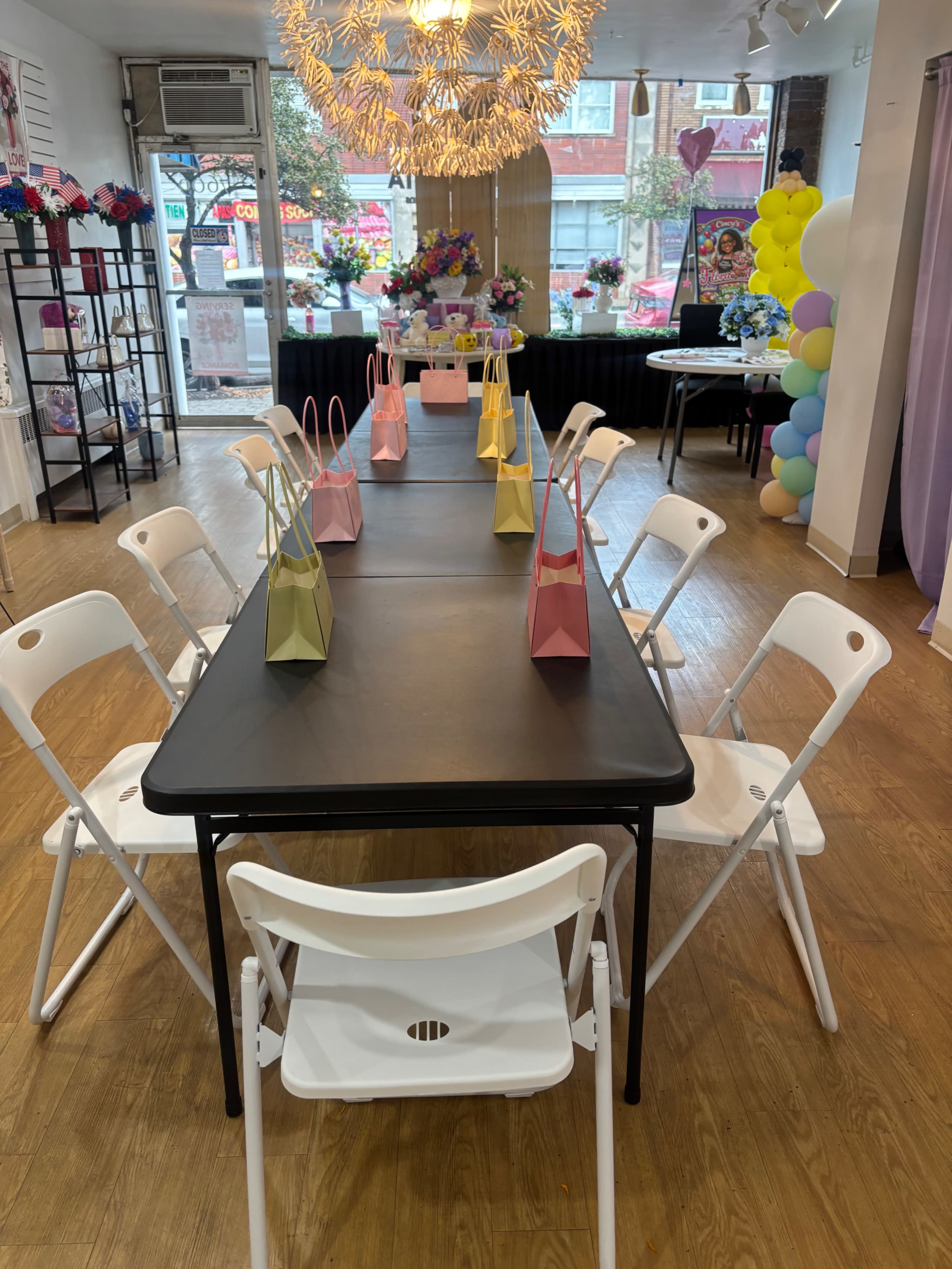 A large table is set with several colored gift bags on each side, surrounded by white folding chairs in a brightly lit room.