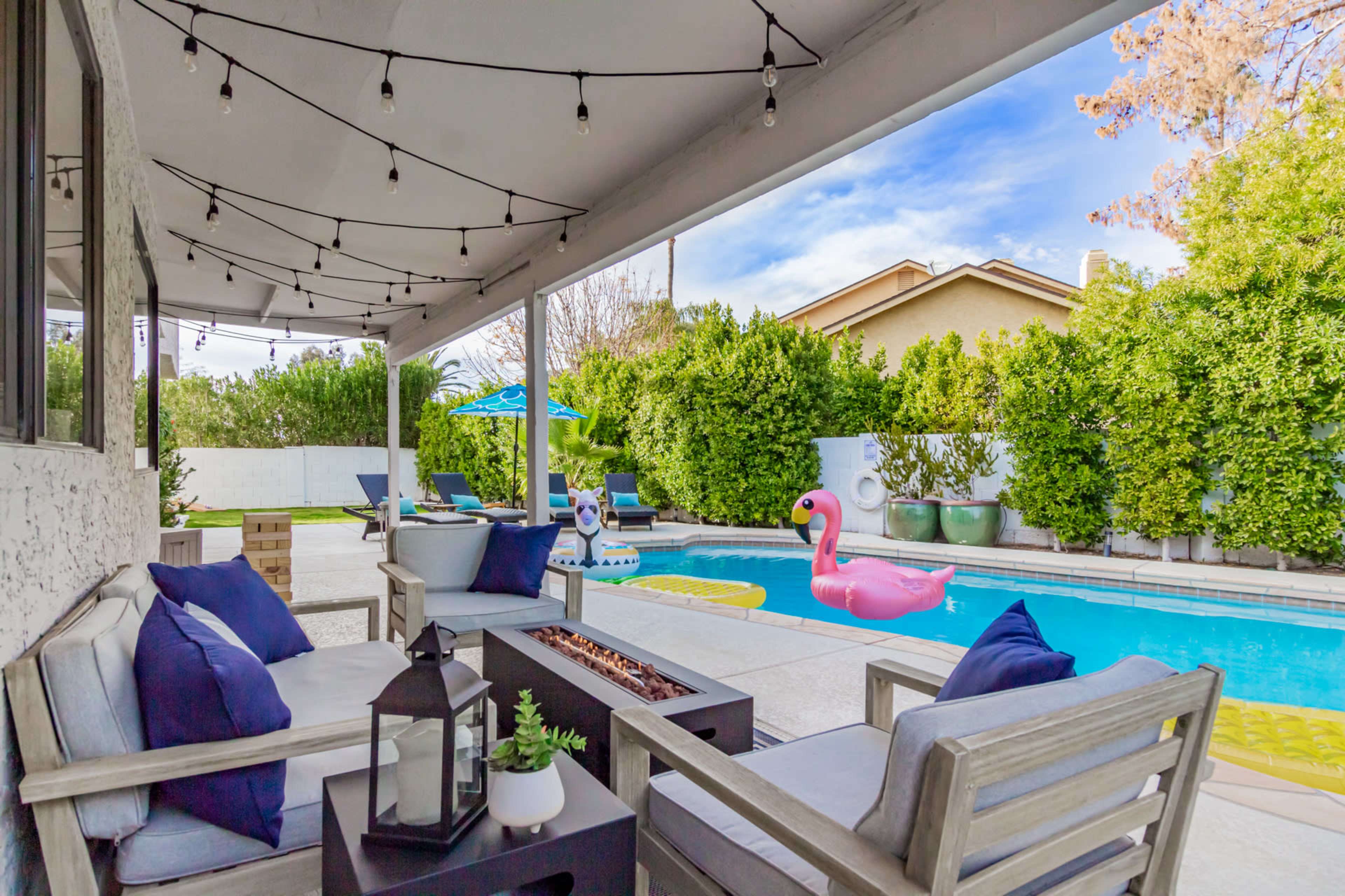A patio with wooden seating and a fire pit overlooks a swimming pool featuring colorful inflatable floaties and surrounded by lush greenery.