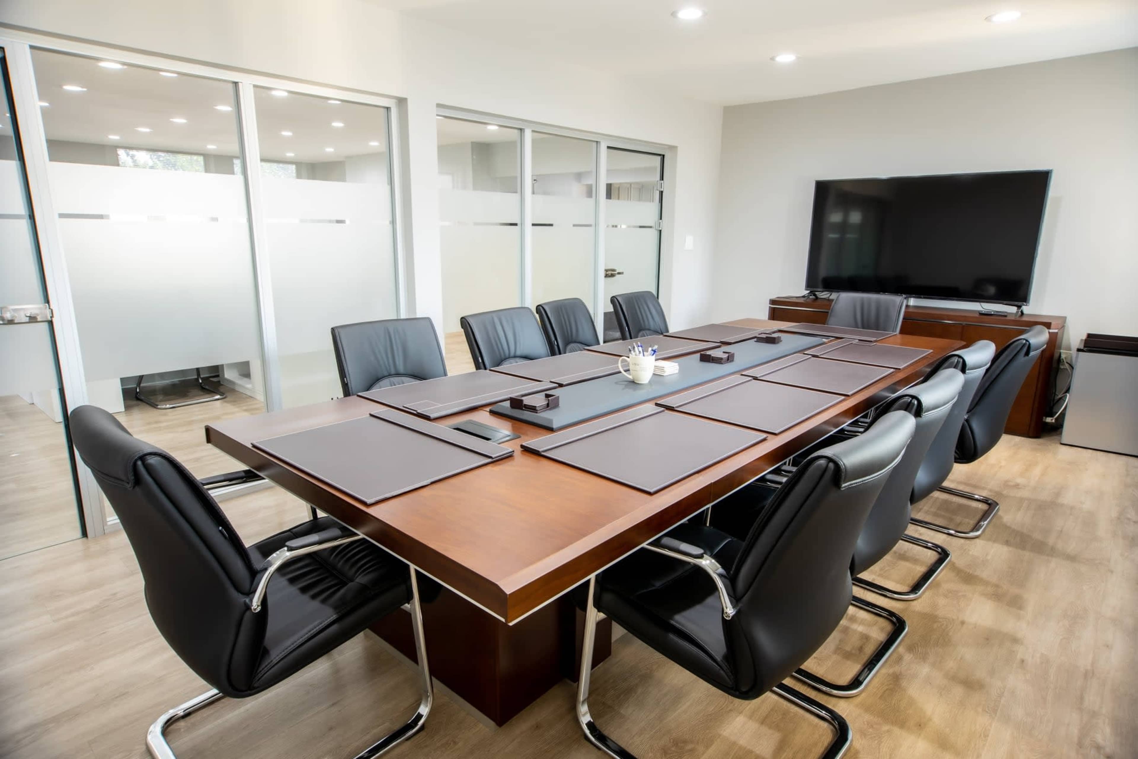 A modern conference room features a large wooden table surrounded by leather chairs, with a television mounted on the wall and glass partitions in the background.