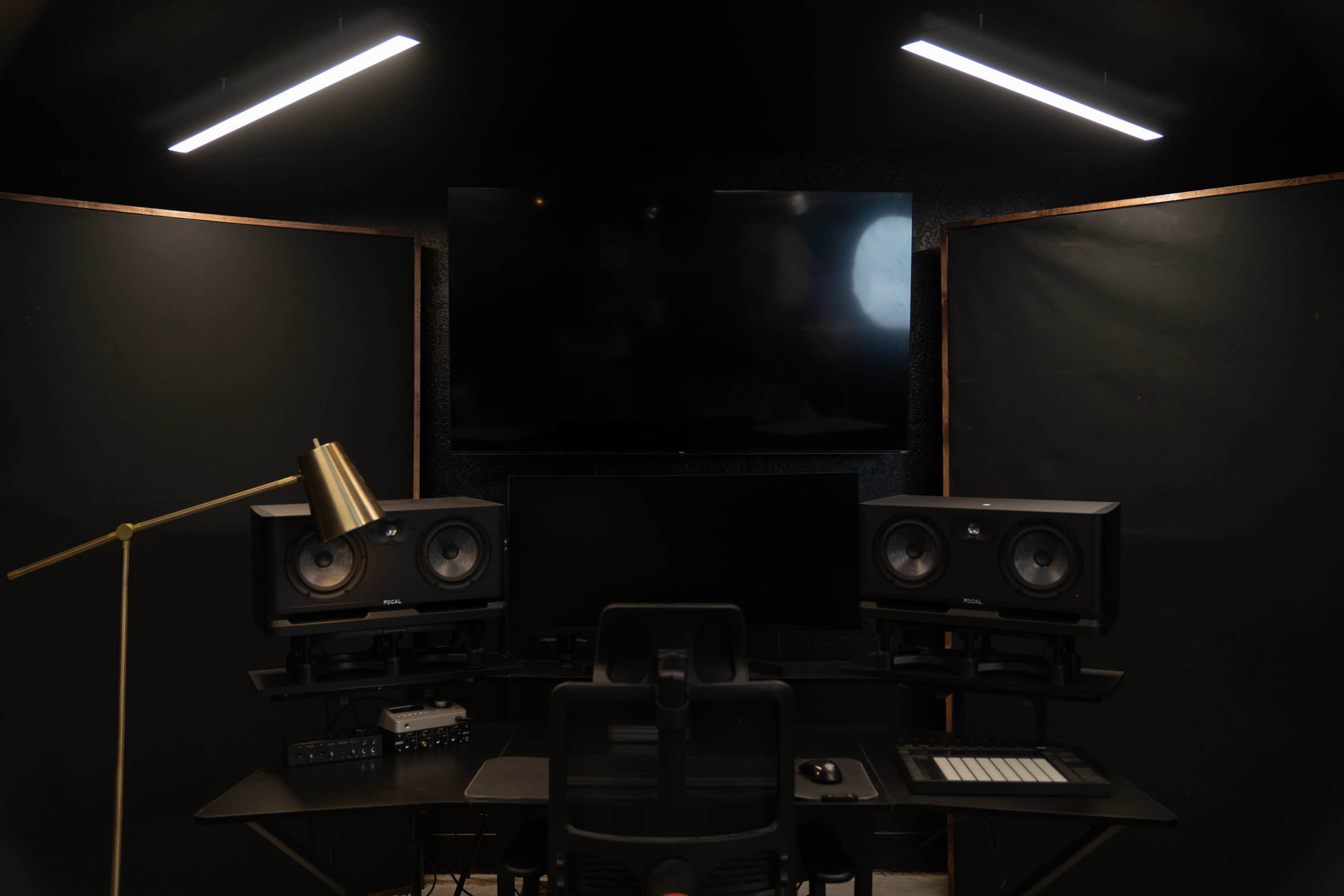 A workstation features dual monitors, studio speakers, and a lamp in a soundproof room with dark walls and overhead lighting.