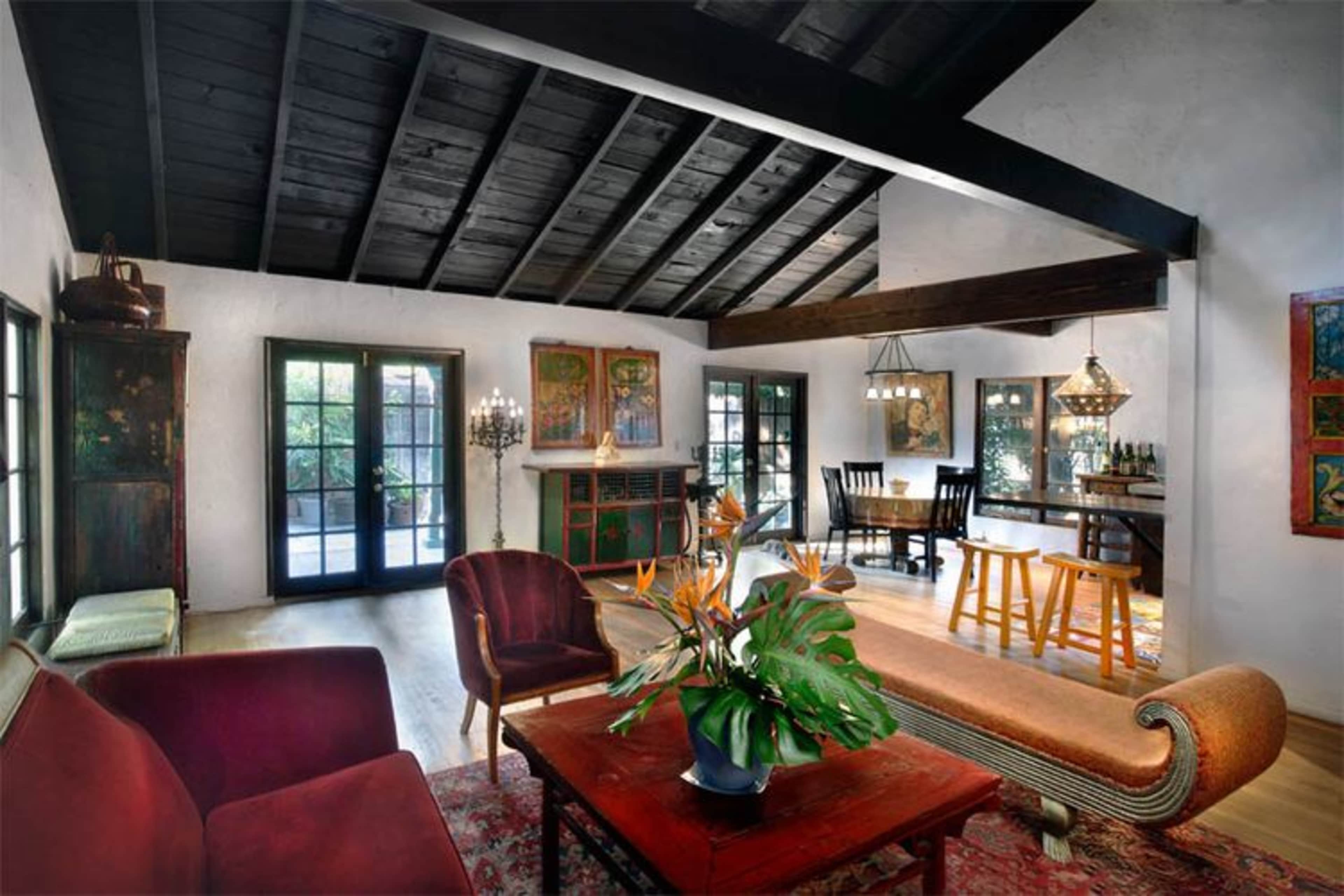 A spacious living room with a high, dark wooden ceiling, featuring a mix of seating areas, decorative plants, and a dining area in the background.
