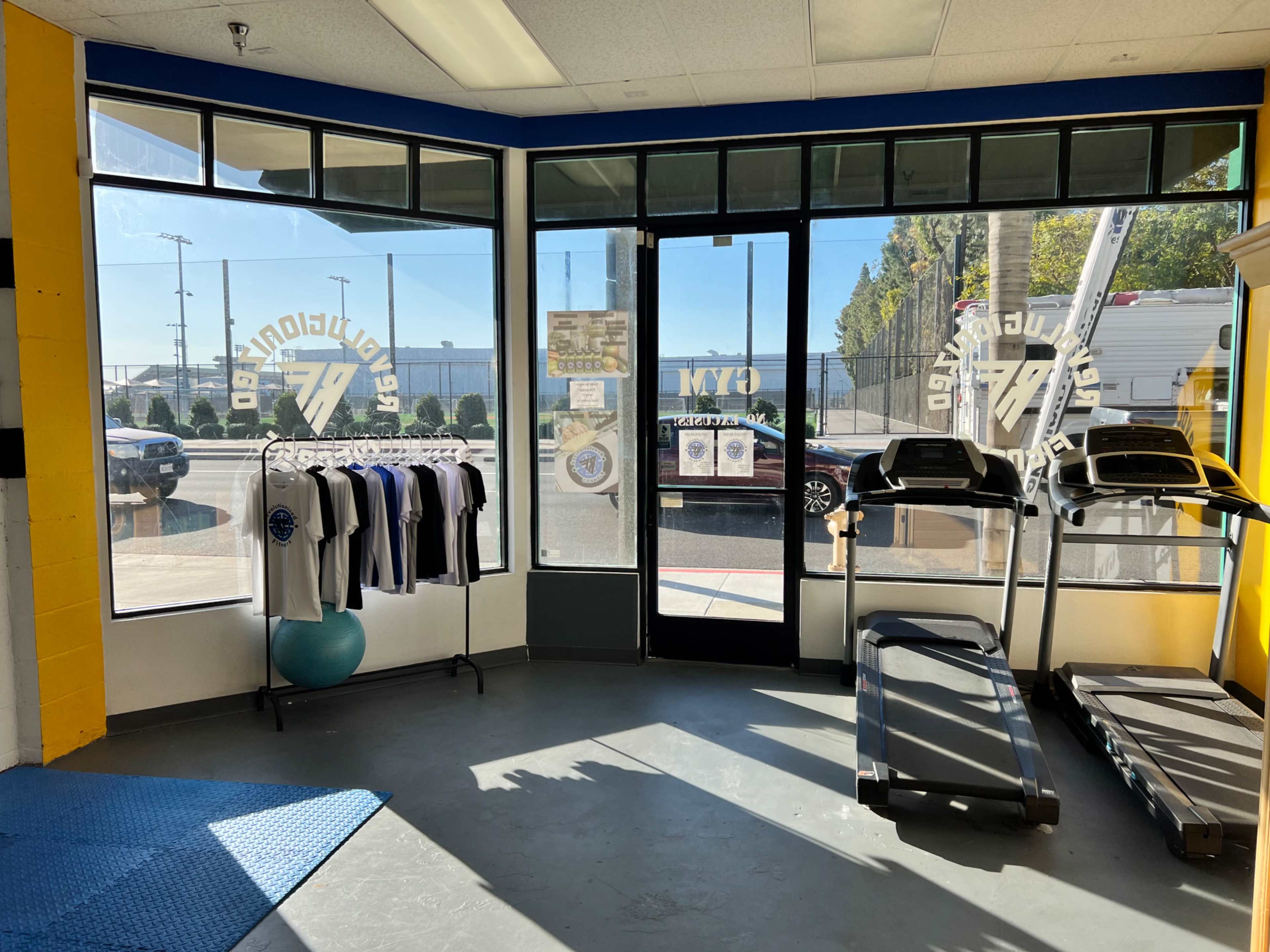 The image shows the interior of a gym, featuring two treadmills, a display of hanging workout shirts, and large windows letting in natural light.