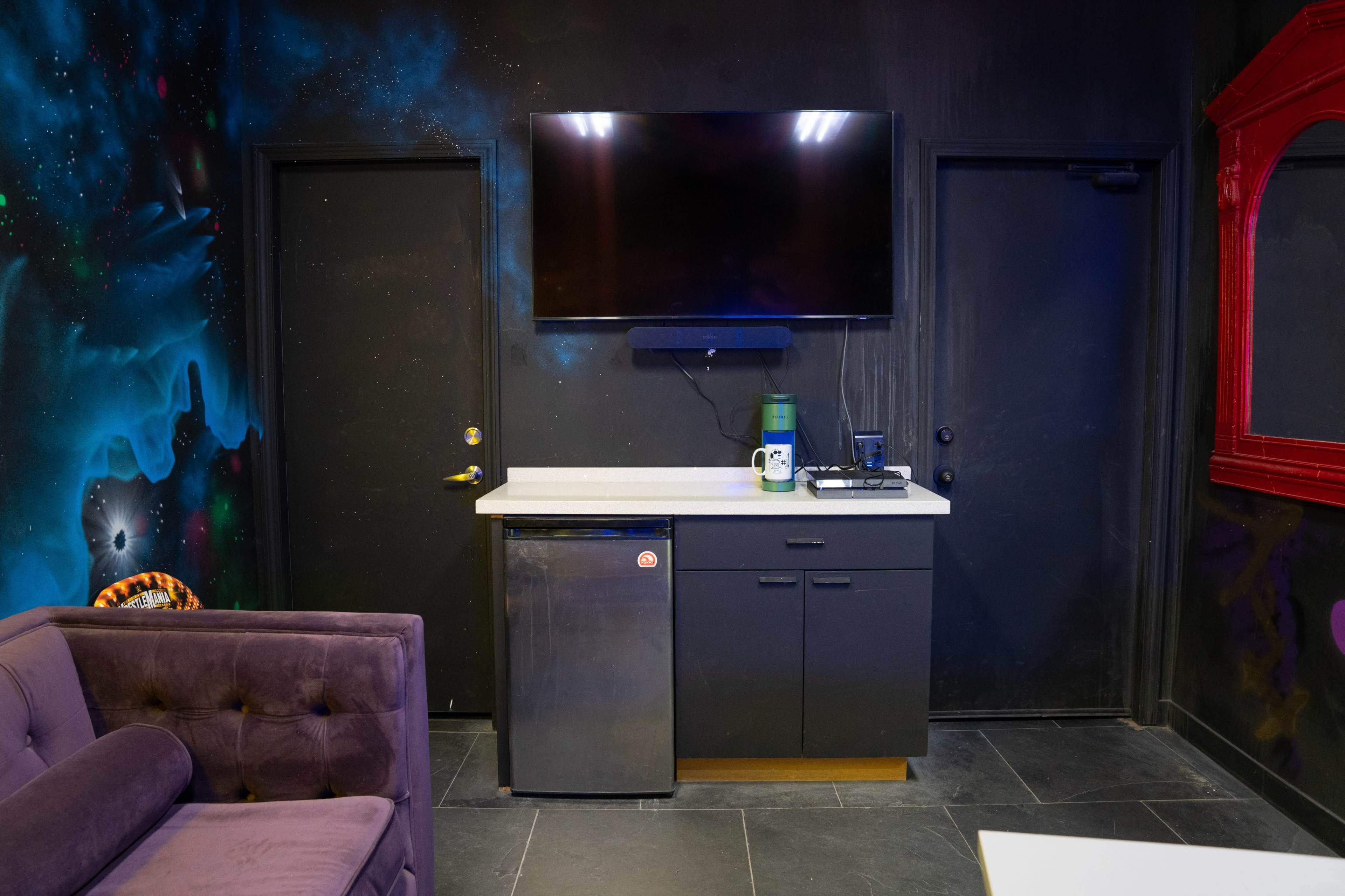 A small lounge area with a dark wall featuring a colorful mural, a mounted TV, a countertop with a mini fridge and other appliances, and a door leading to another room.