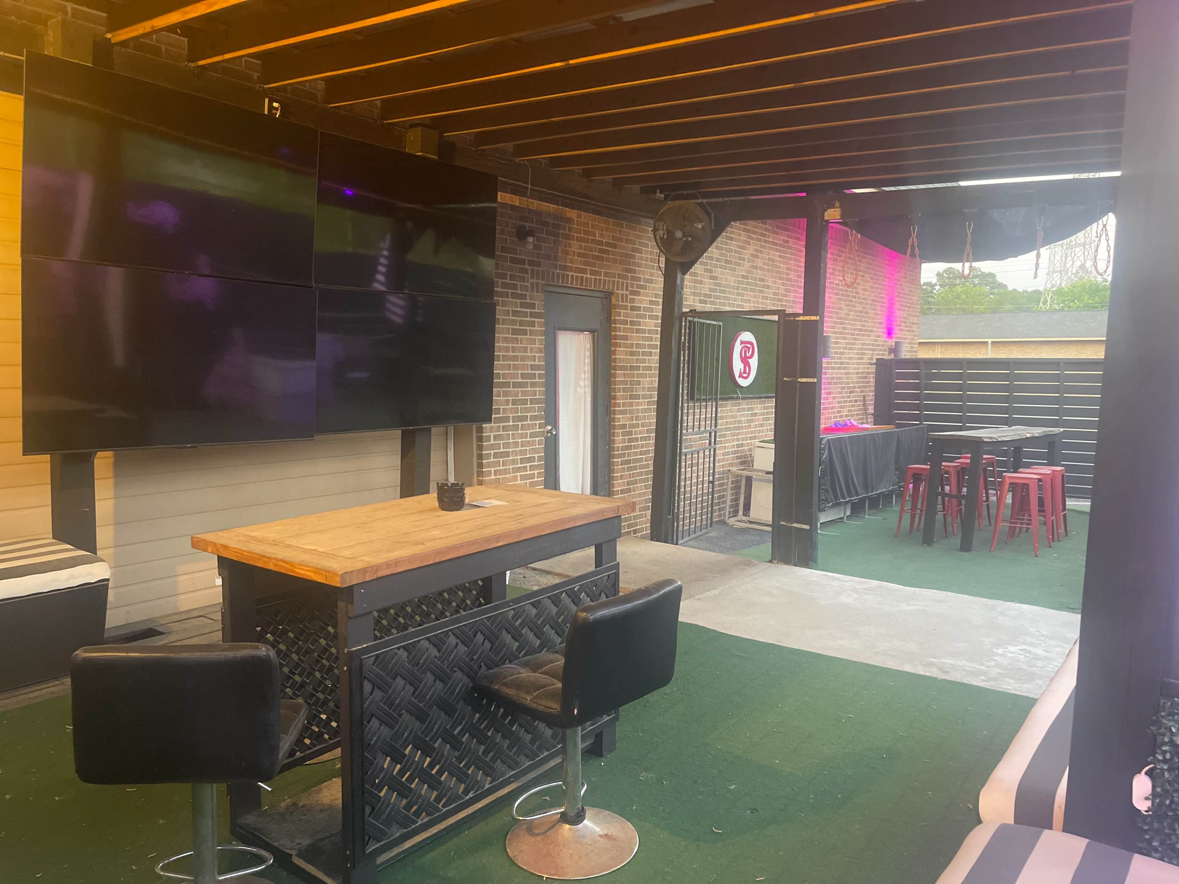 The image shows an outdoor bar area with a wooden table, black chairs, several large screens mounted on the wall, and a fenced section leading to another area.