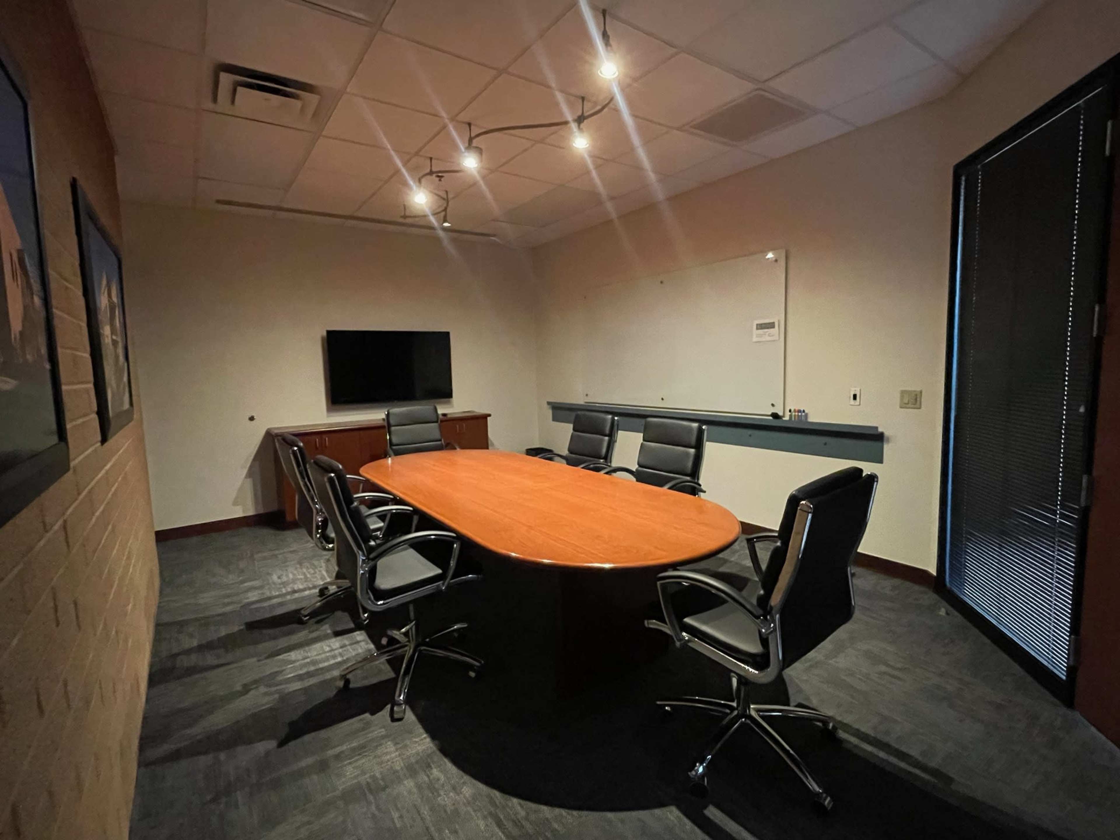 A conference room features a large oval table surrounded by black chairs, with a television on the wall and a whiteboard at the front.