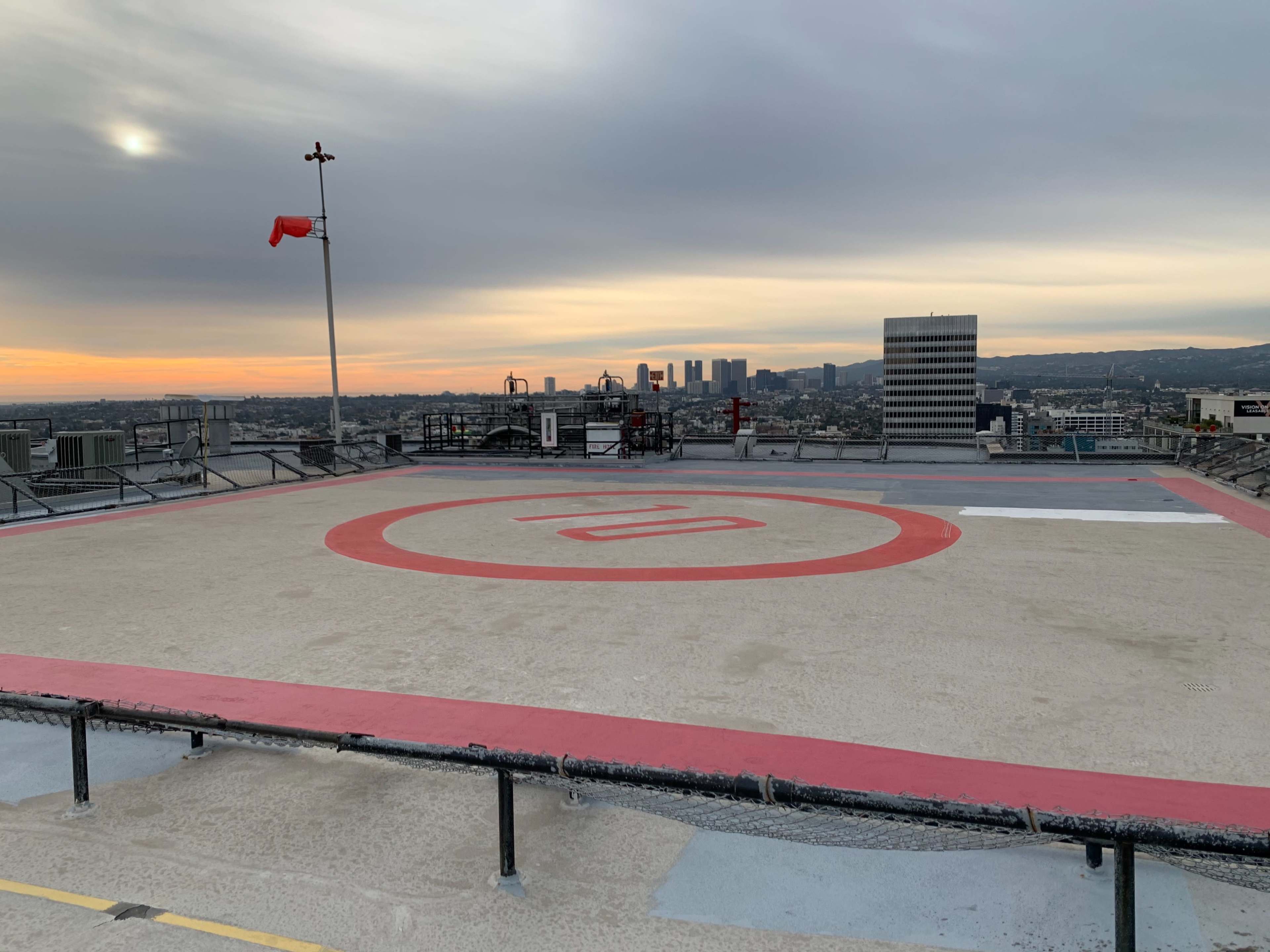 A helicopter landing pad is situated on a rooftop, with a sunset view of the city skyline in the background.