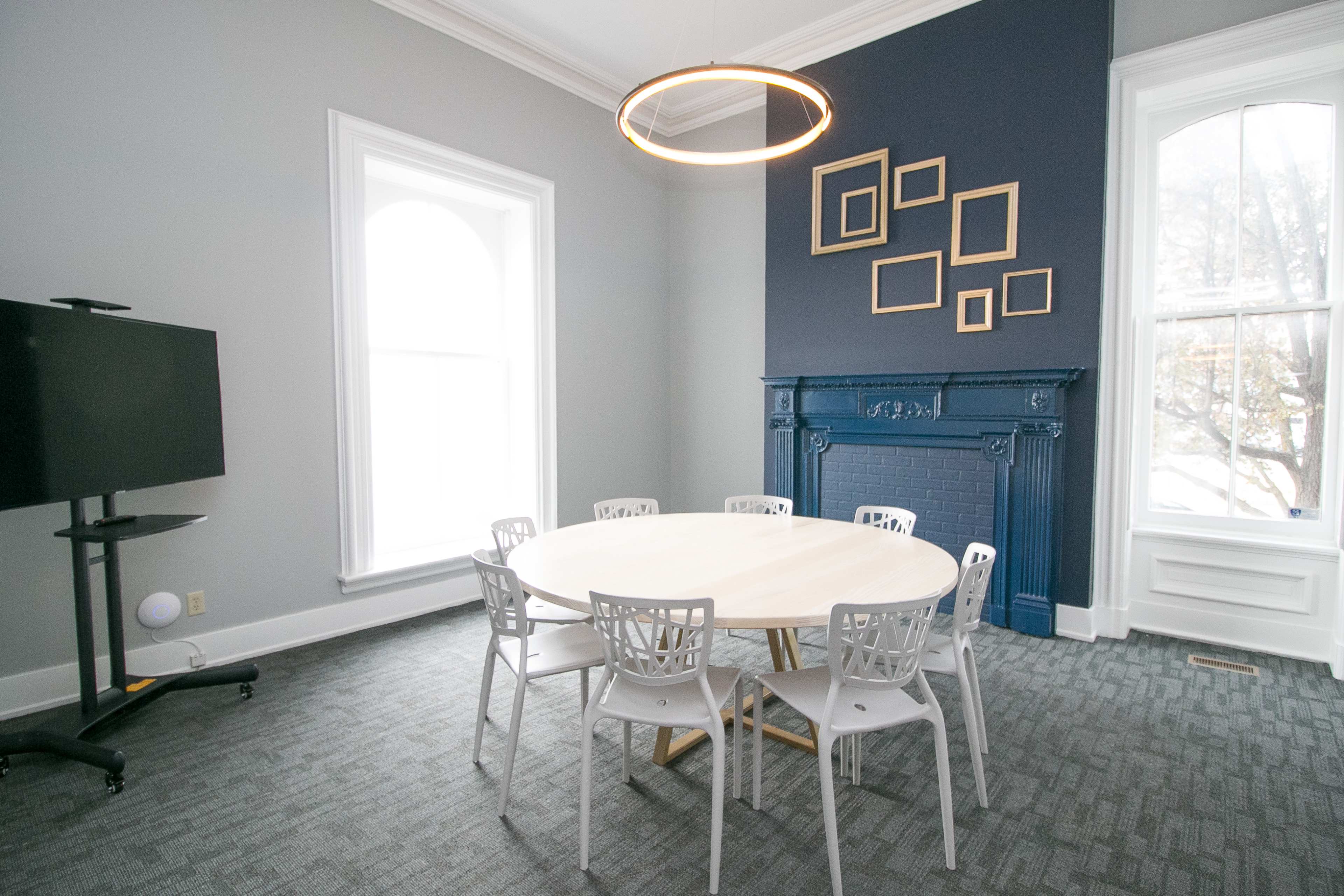A round wooden table is surrounded by six white chairs in a light-filled room featuring a dark blue wall with framed pictures and a decorative fireplace.