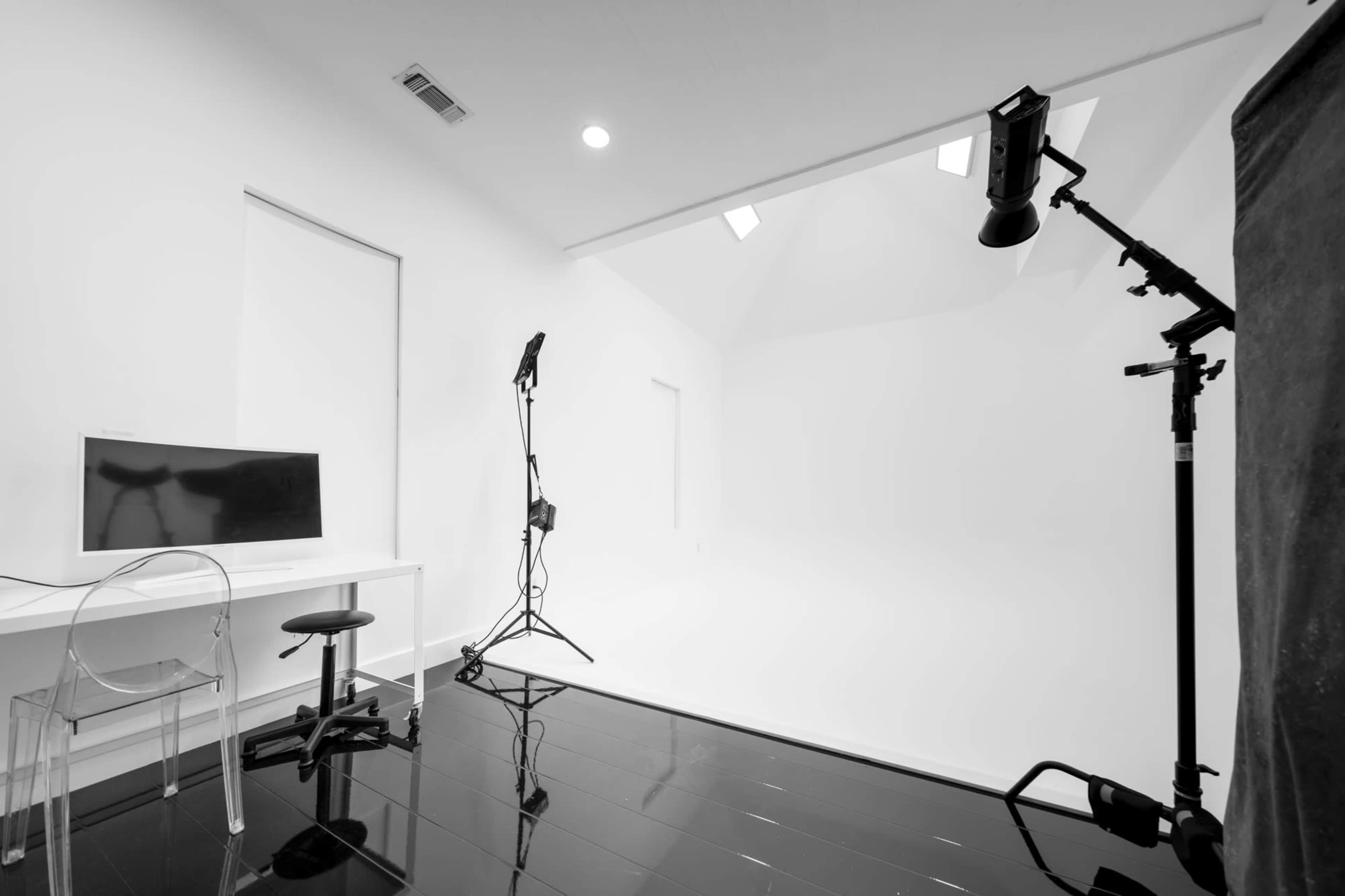 A minimalist photography studio features a white backdrop, a computer desk, and lighting equipment on stands.