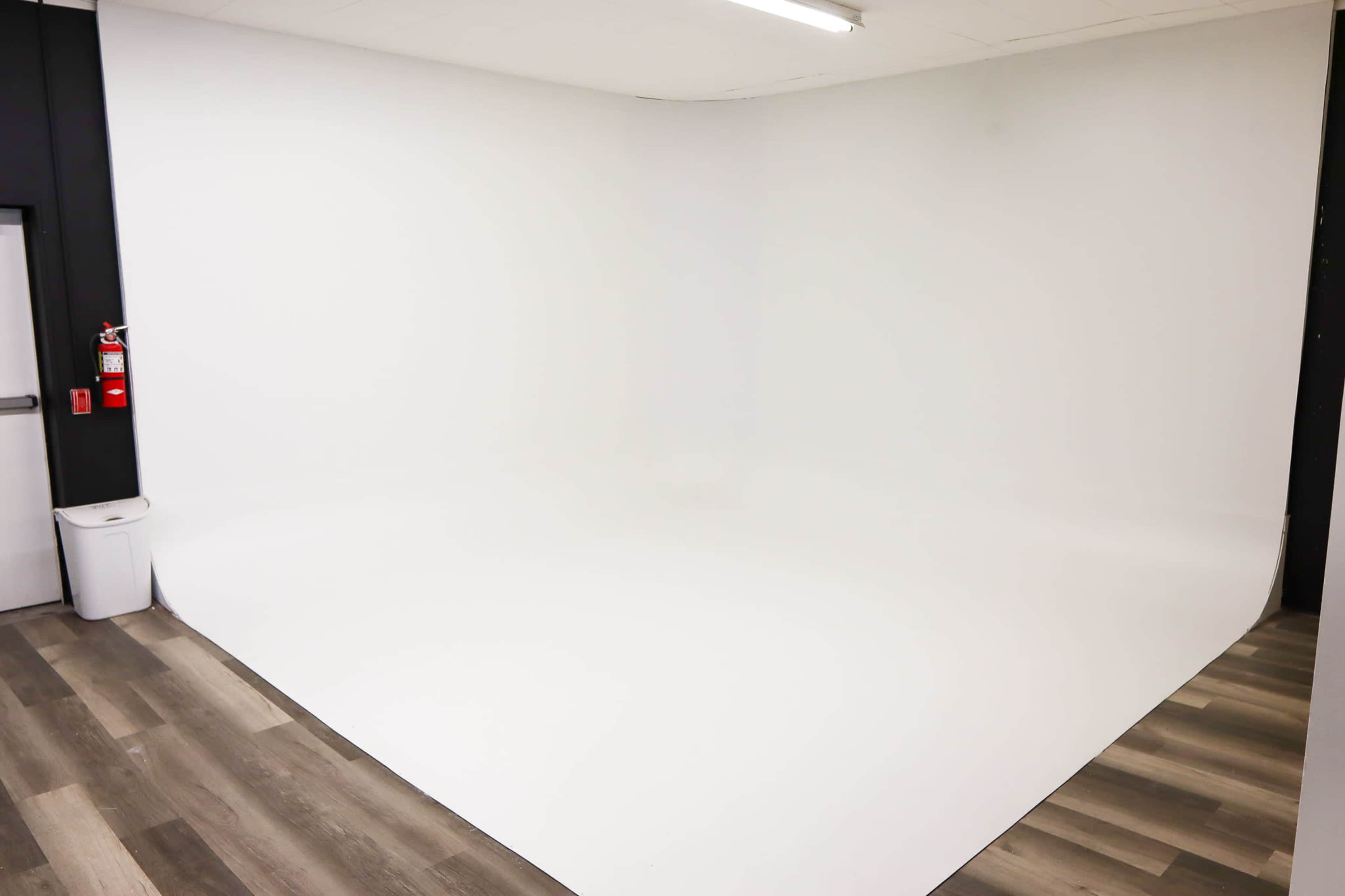 The image shows a spacious, empty room with a seamless white backdrop and wooden flooring.
