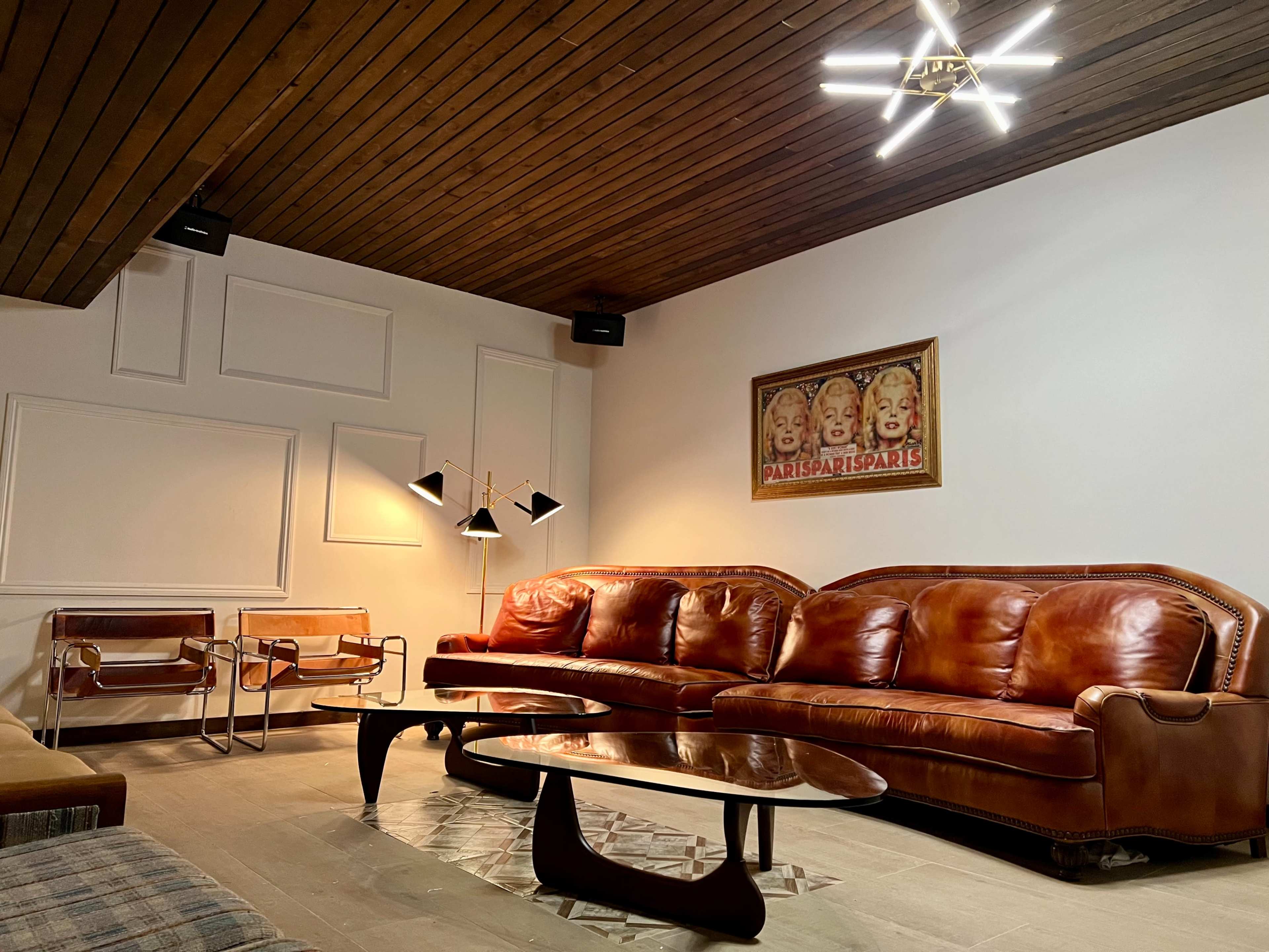 A cozy living room featuring a brown leather sofa, a mid-century modern chair, and a distinctive ceiling light, with a vintage Parisian poster on the wall.