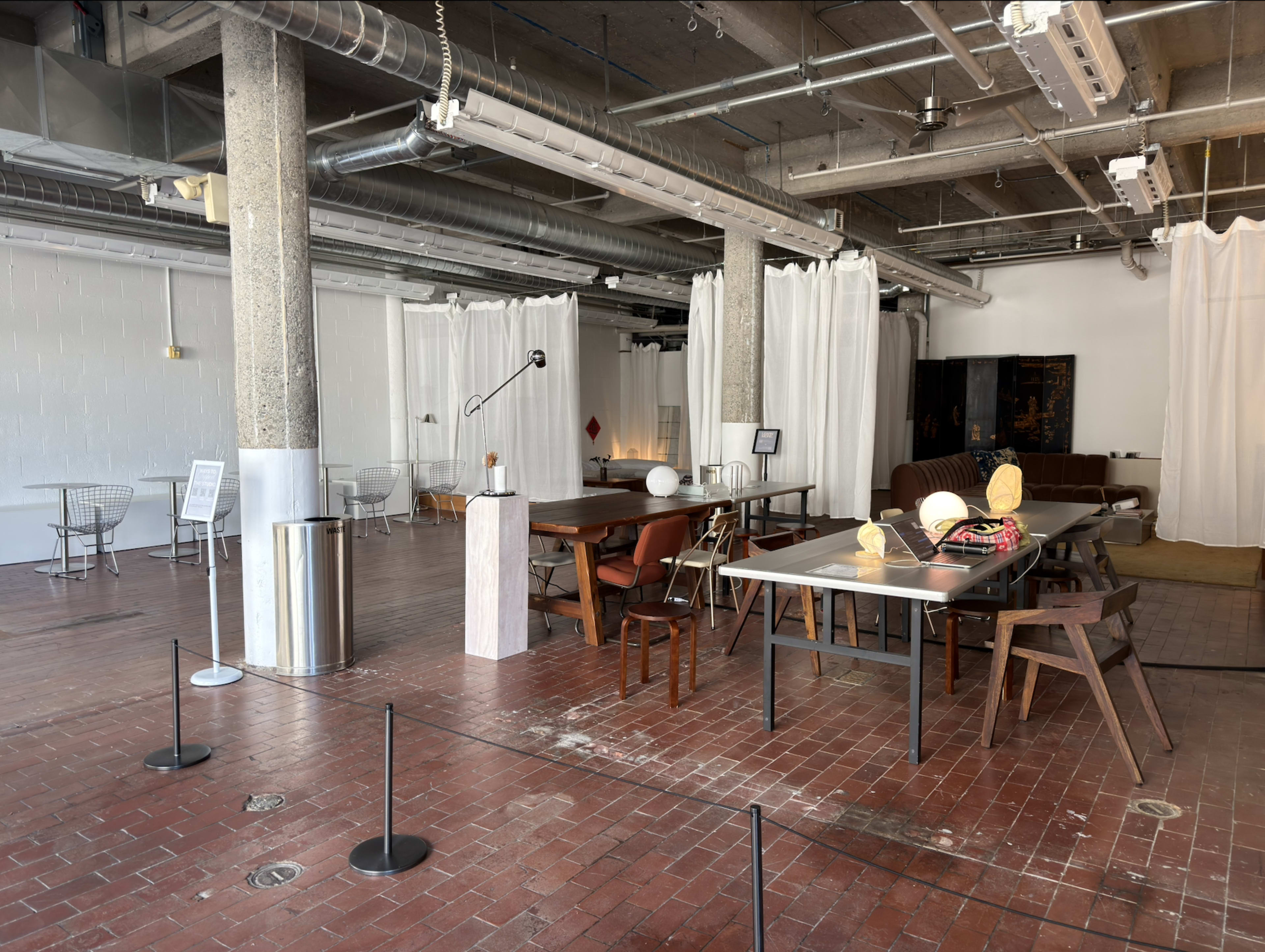 An industrial-style space features various tables and seating arranged throughout, with white curtains dividing areas and exposed ductwork above.