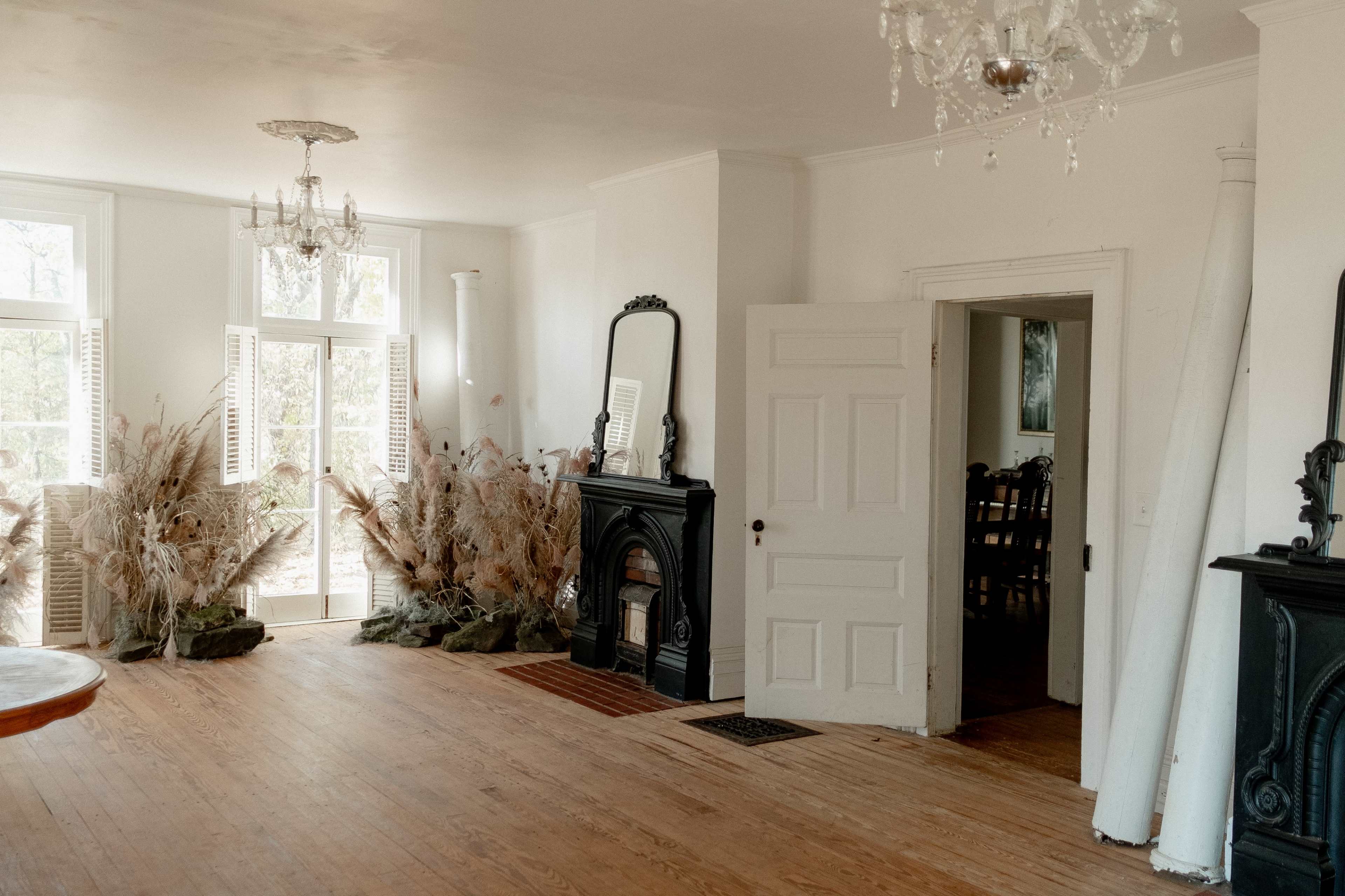 A spacious room with wooden floors, a large door leading to an outdoor area, and decorative elements including a chandelier and arrangements of dried plants by a fireplace.