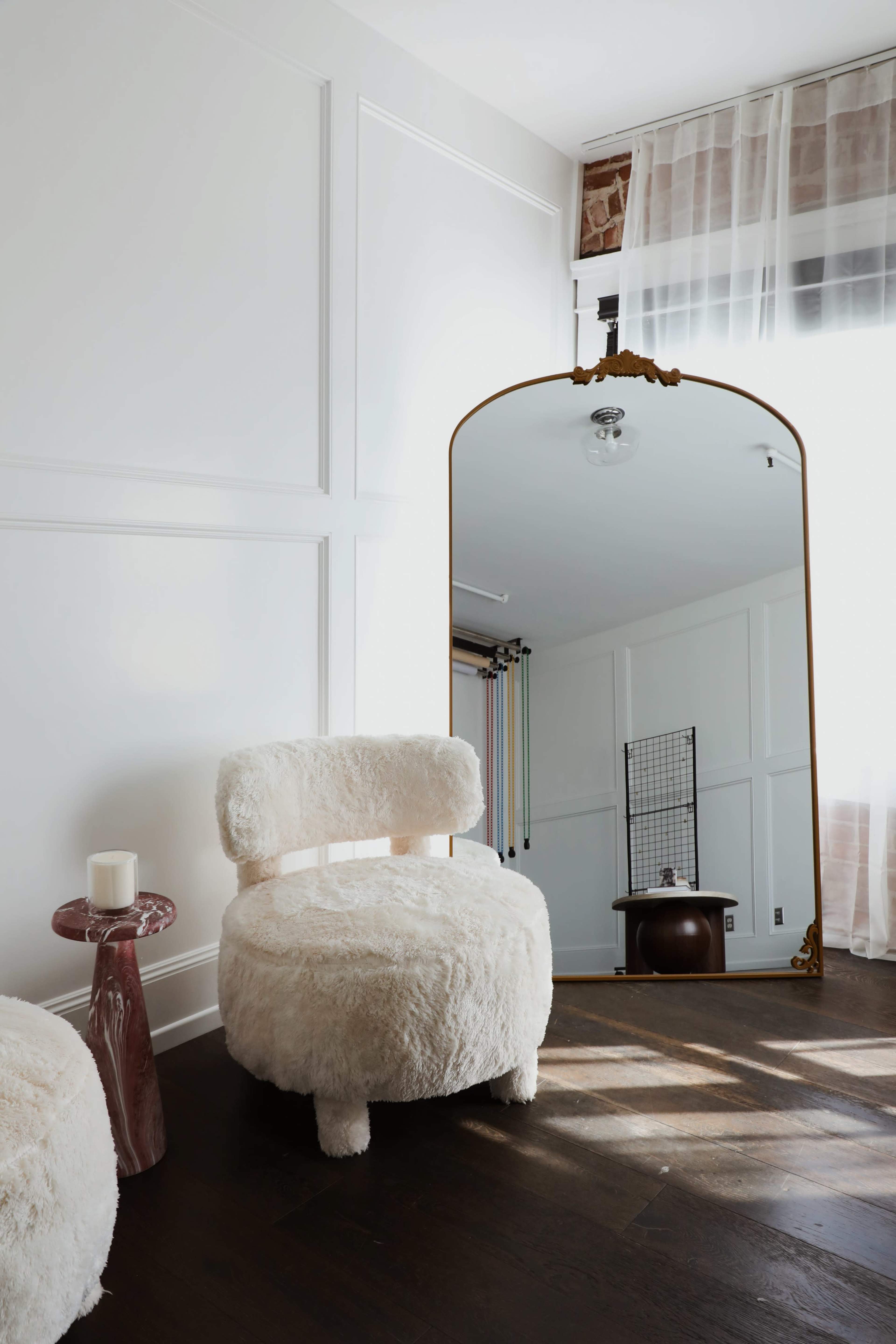 The image shows a cozy interior featuring a fluffy white chair, a small table with a candle, and a large mirror reflecting the room.