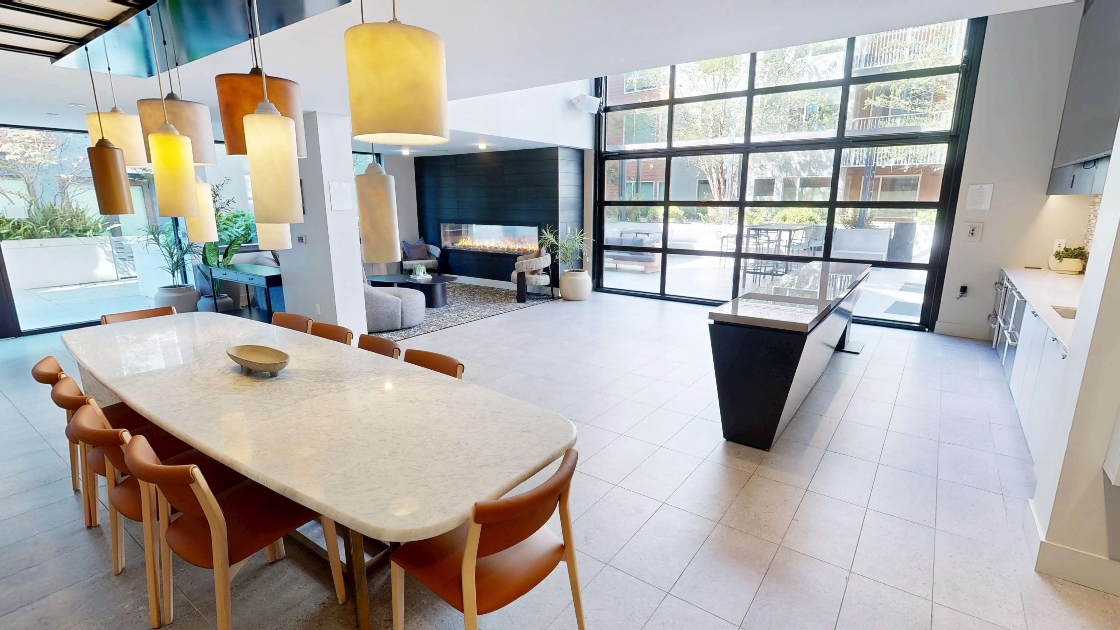 The image shows a modern dining area with a large marble table and leather chairs, adjacent to a bright living space featuring large glass windows, a fireplace, and greenery.