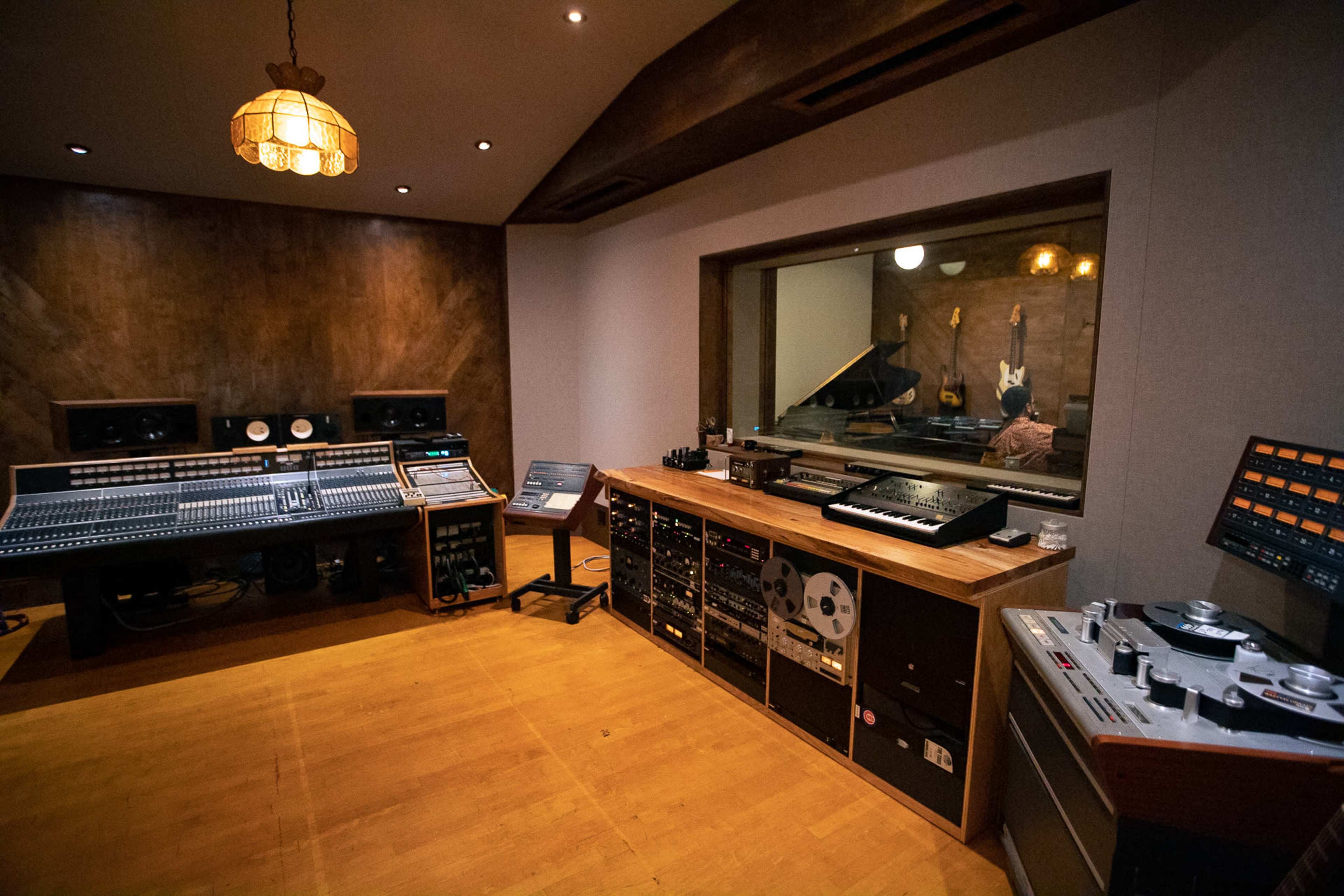 A recording studio with sound equipment, a control desk, and a glass partition overlooking a piano.