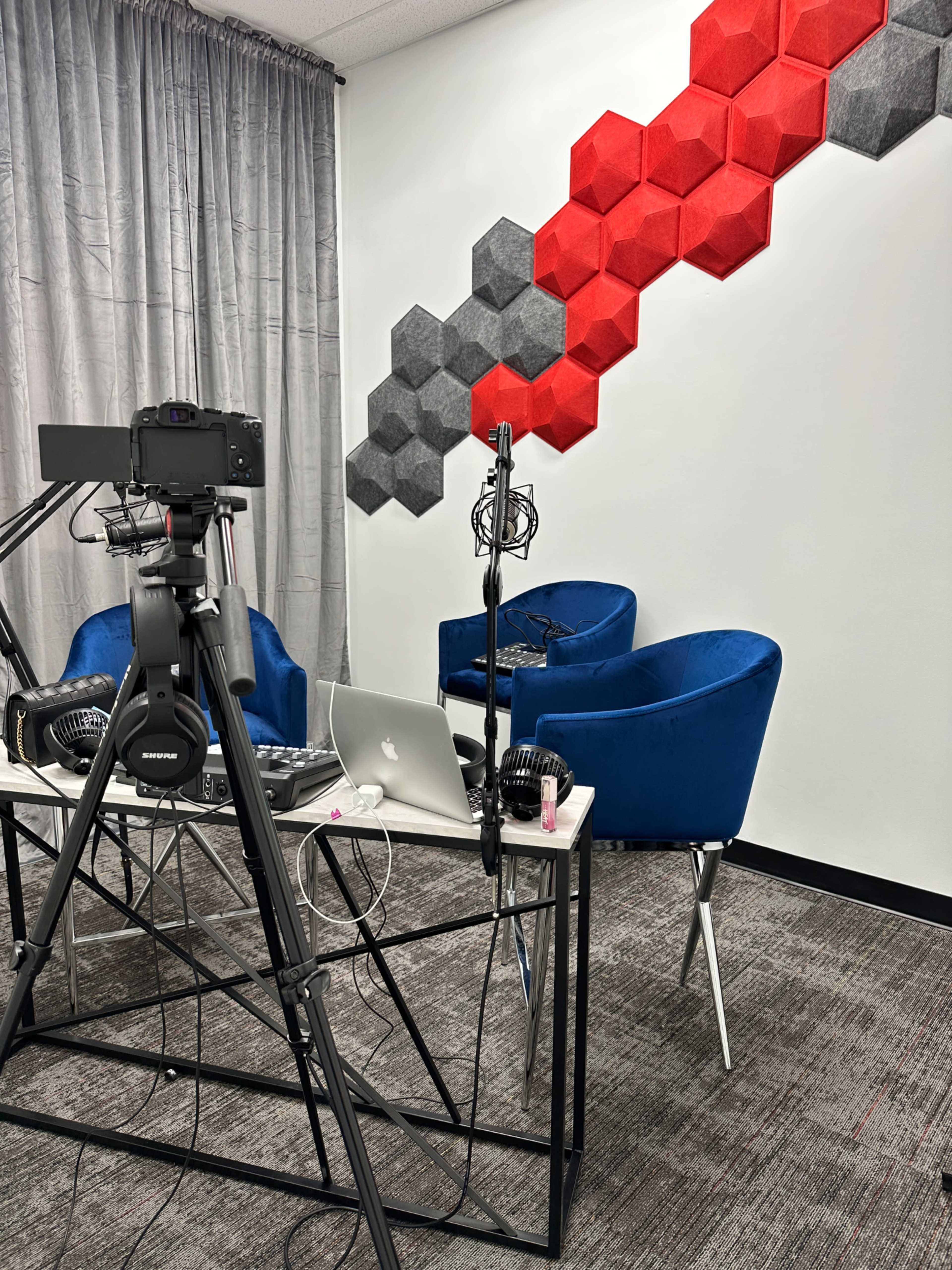 A recording studio setup with two blue chairs, a table holding a laptop, and a microphone, alongside a wall decorated with red and gray hexagonal panels.