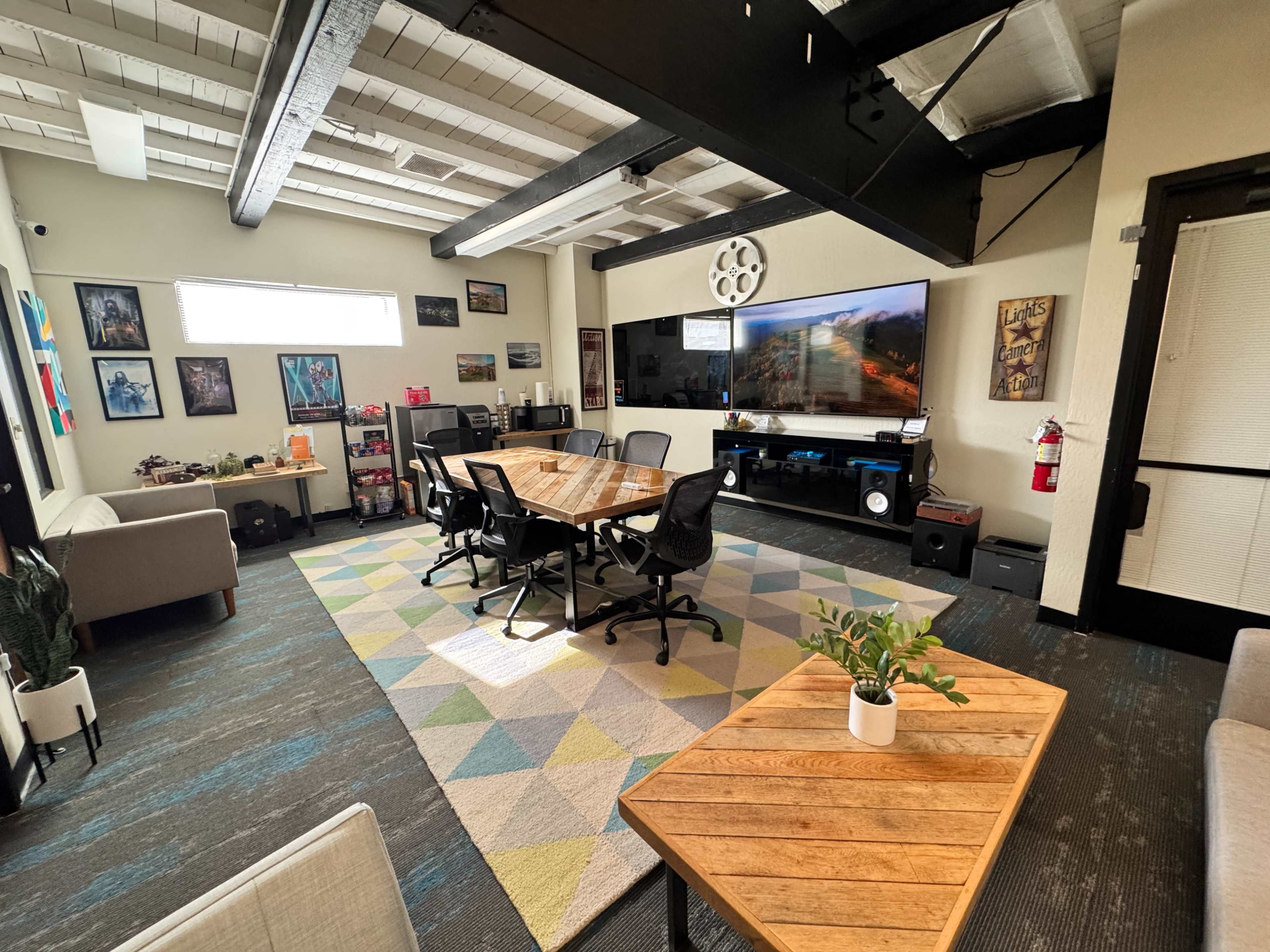 The image shows a modern office meeting room with a large wooden table surrounded by black chairs, a TV mounted on the wall, and decorative artwork.