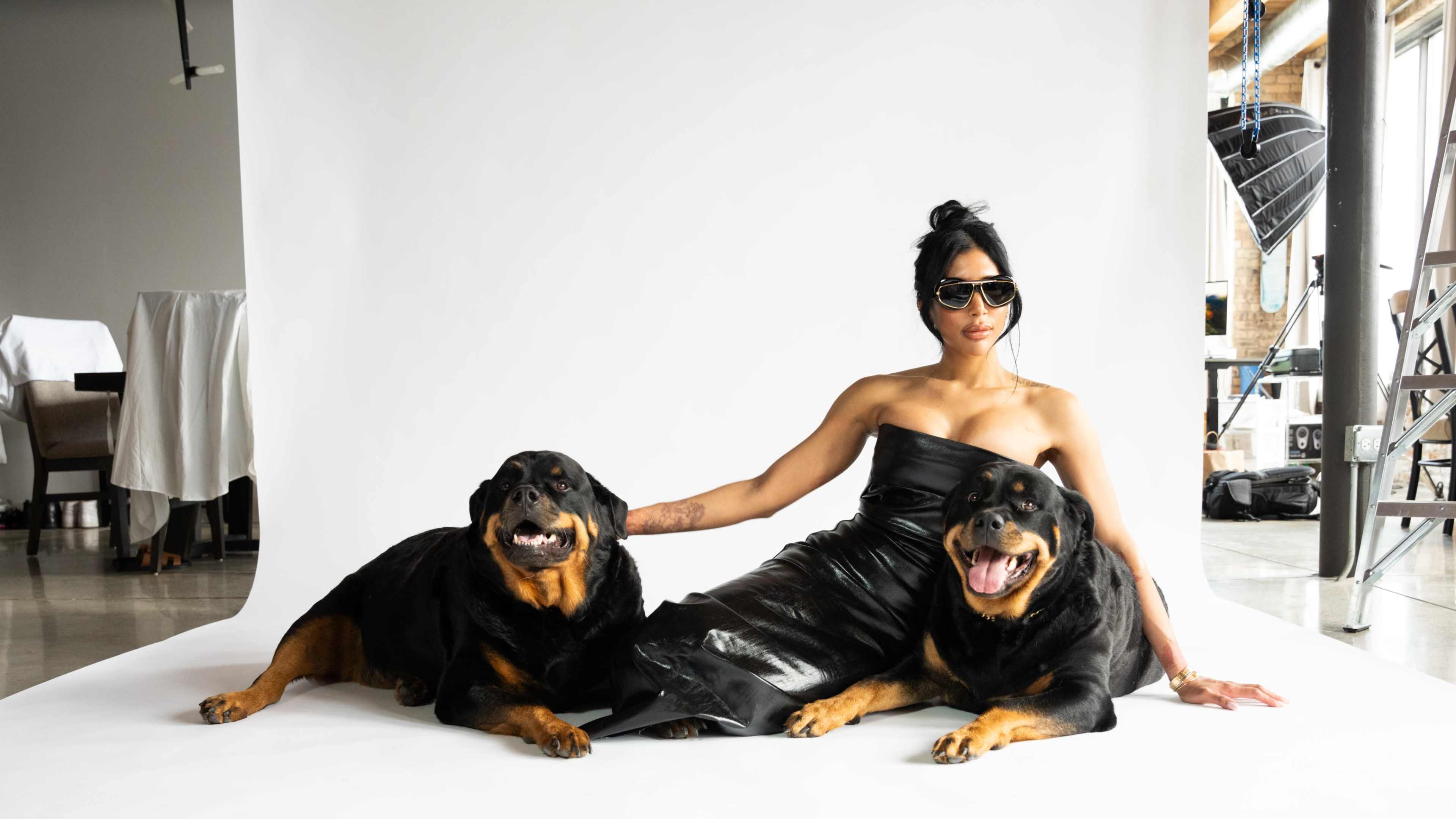 A woman wearing sunglasses sits on a white backdrop with two Rottweiler dogs beside her.