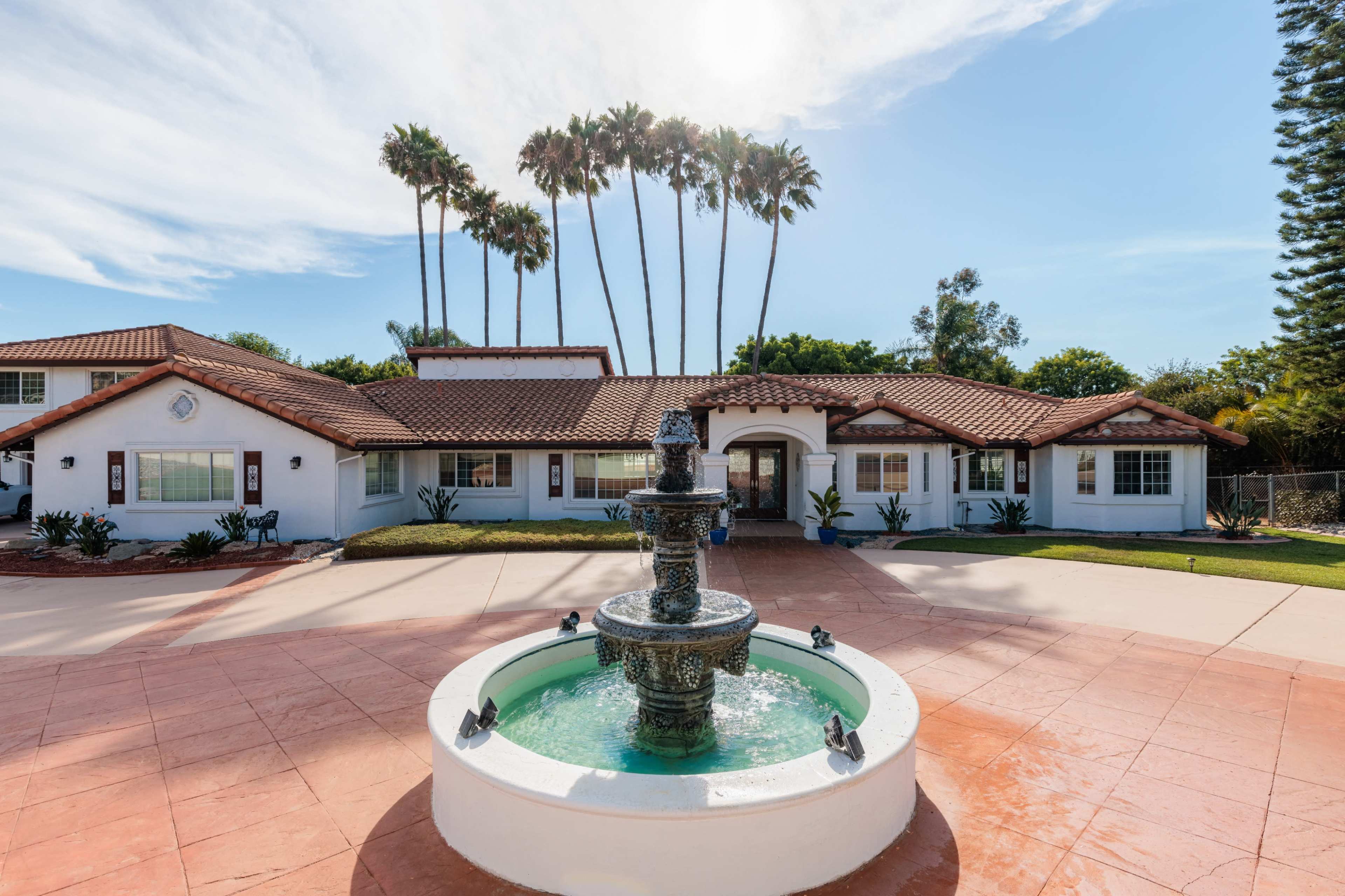 A fountain is centered in a landscaped courtyard in front of a large, single-story house with a red-tiled roof and palm trees in the background.
