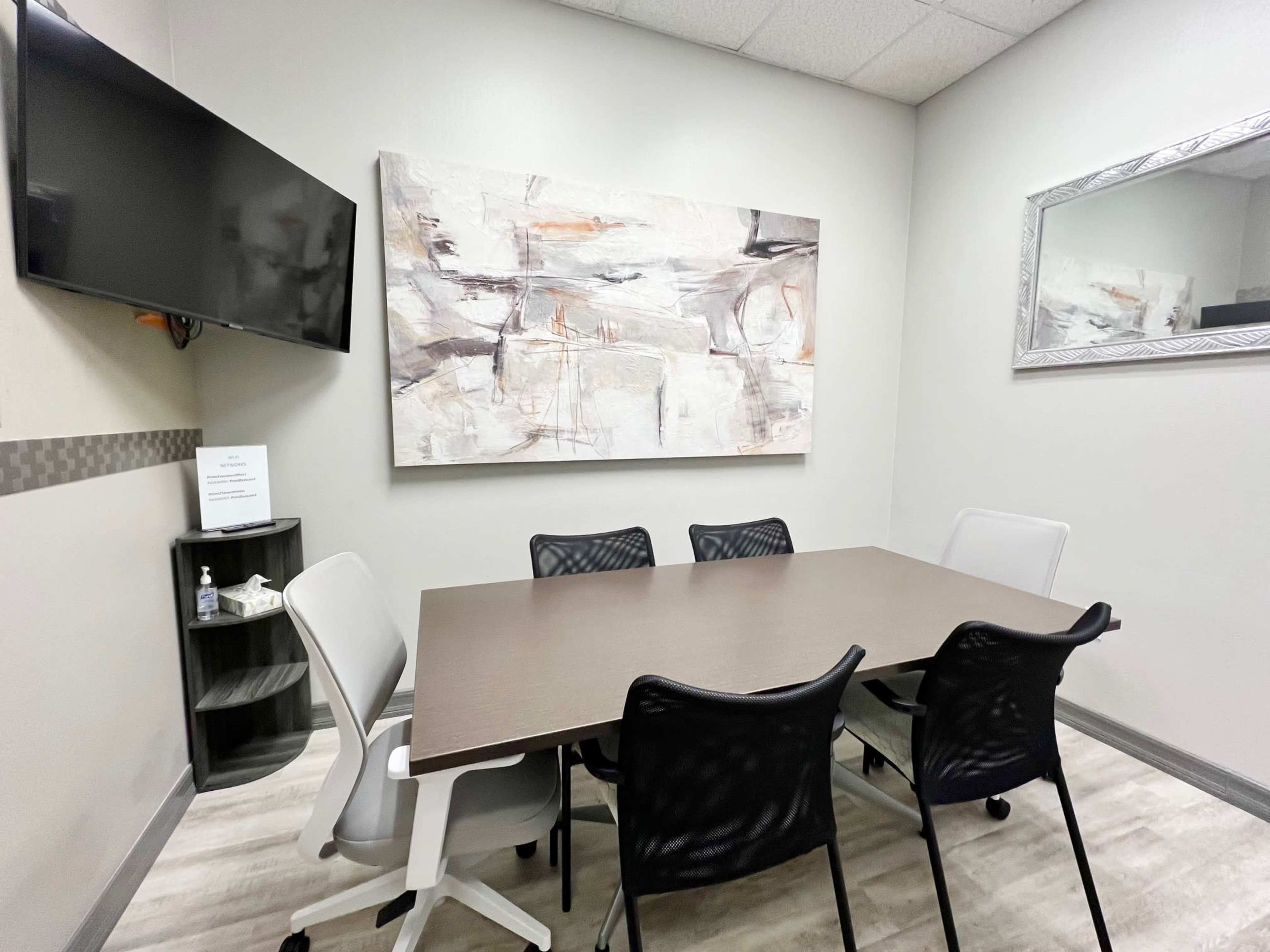 The image shows a small meeting room with a rectangular table surrounded by six chairs, a wall-mounted TV, and abstract artwork on the wall.