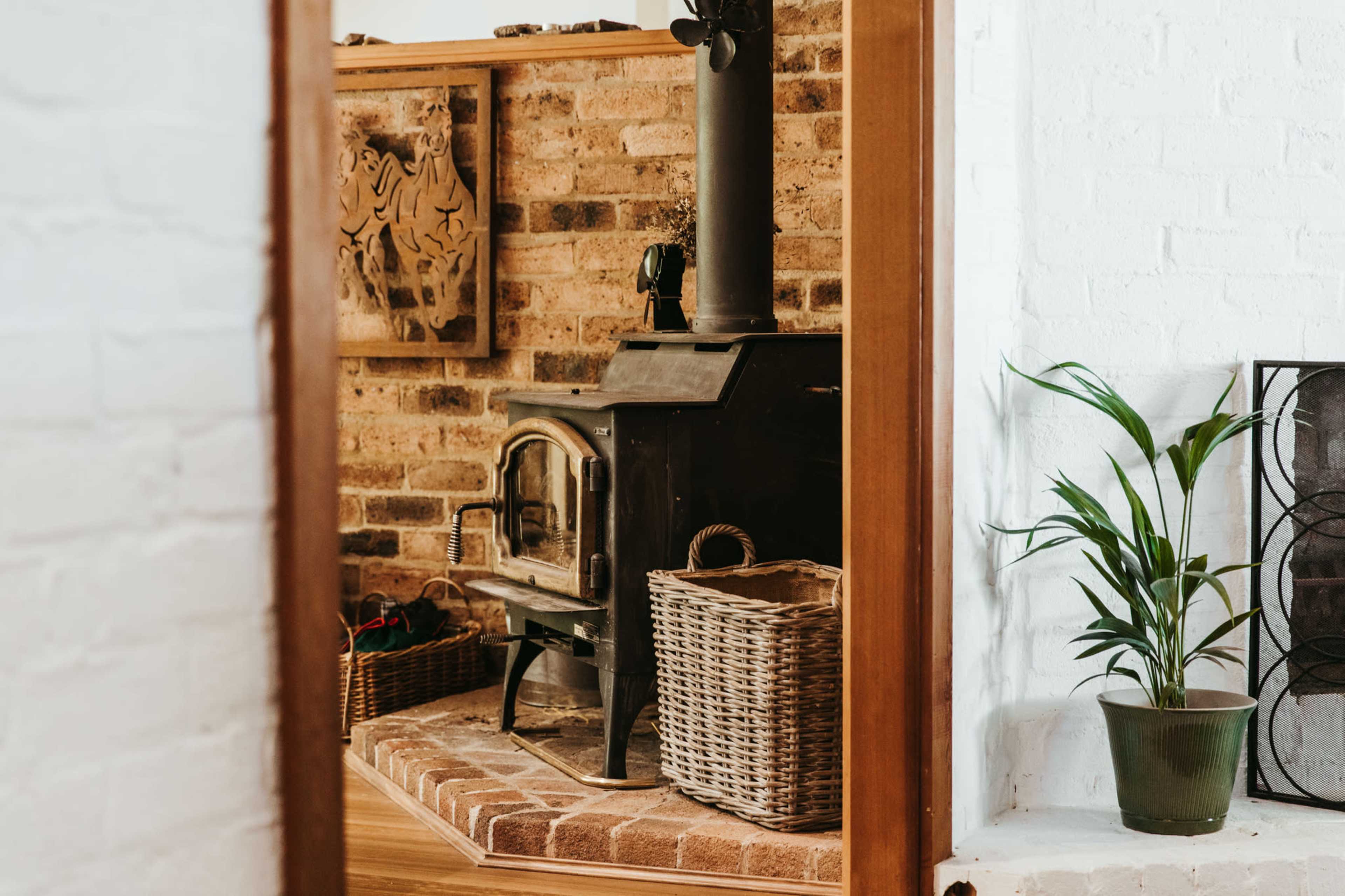 A cozy interior with a brick fireplace, wood-burning stove, woven baskets, and a potted plant.