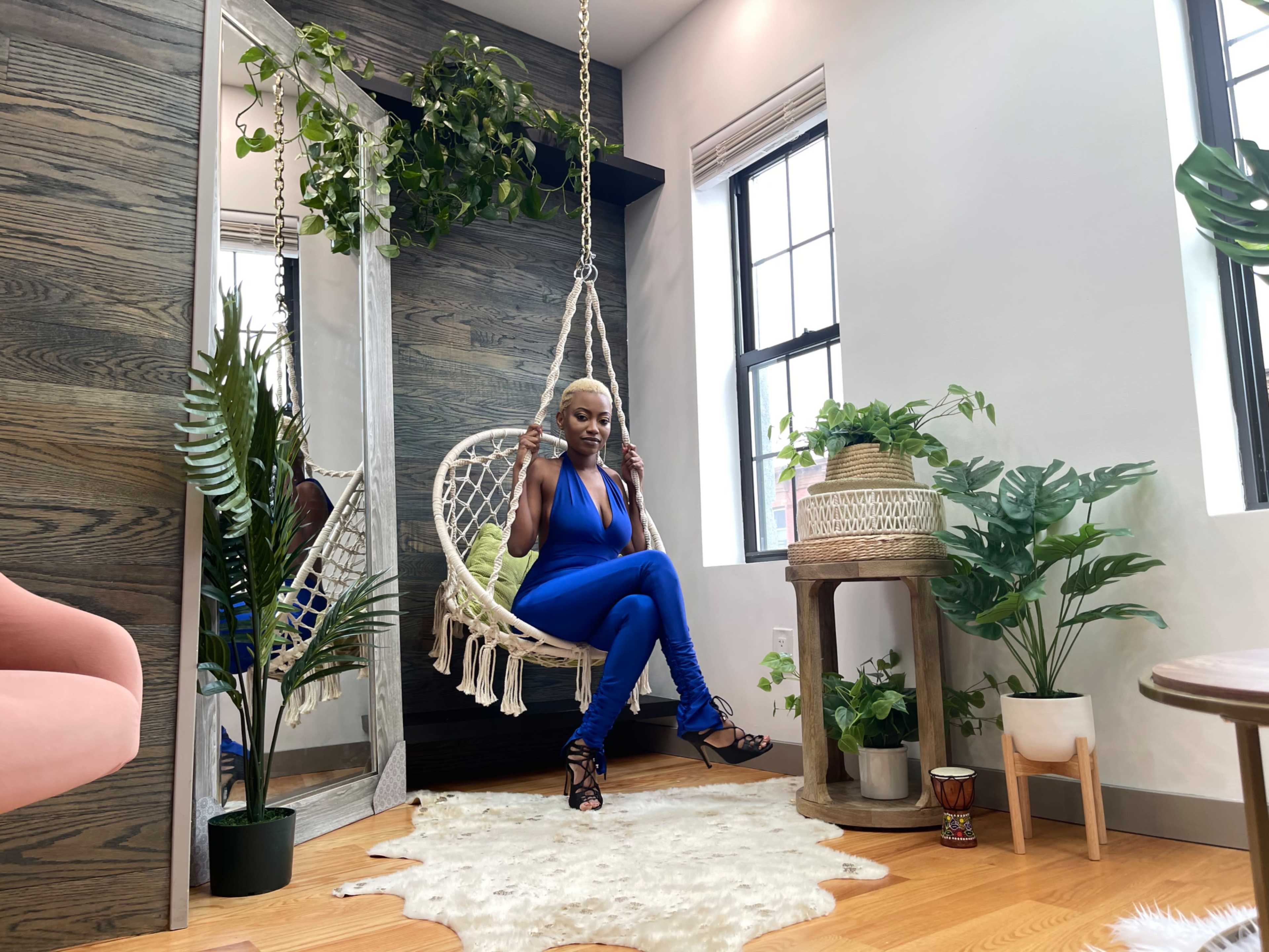 A person sits on a hanging macramé chair surrounded by various indoor plants in a well-lit room with wooden accents.