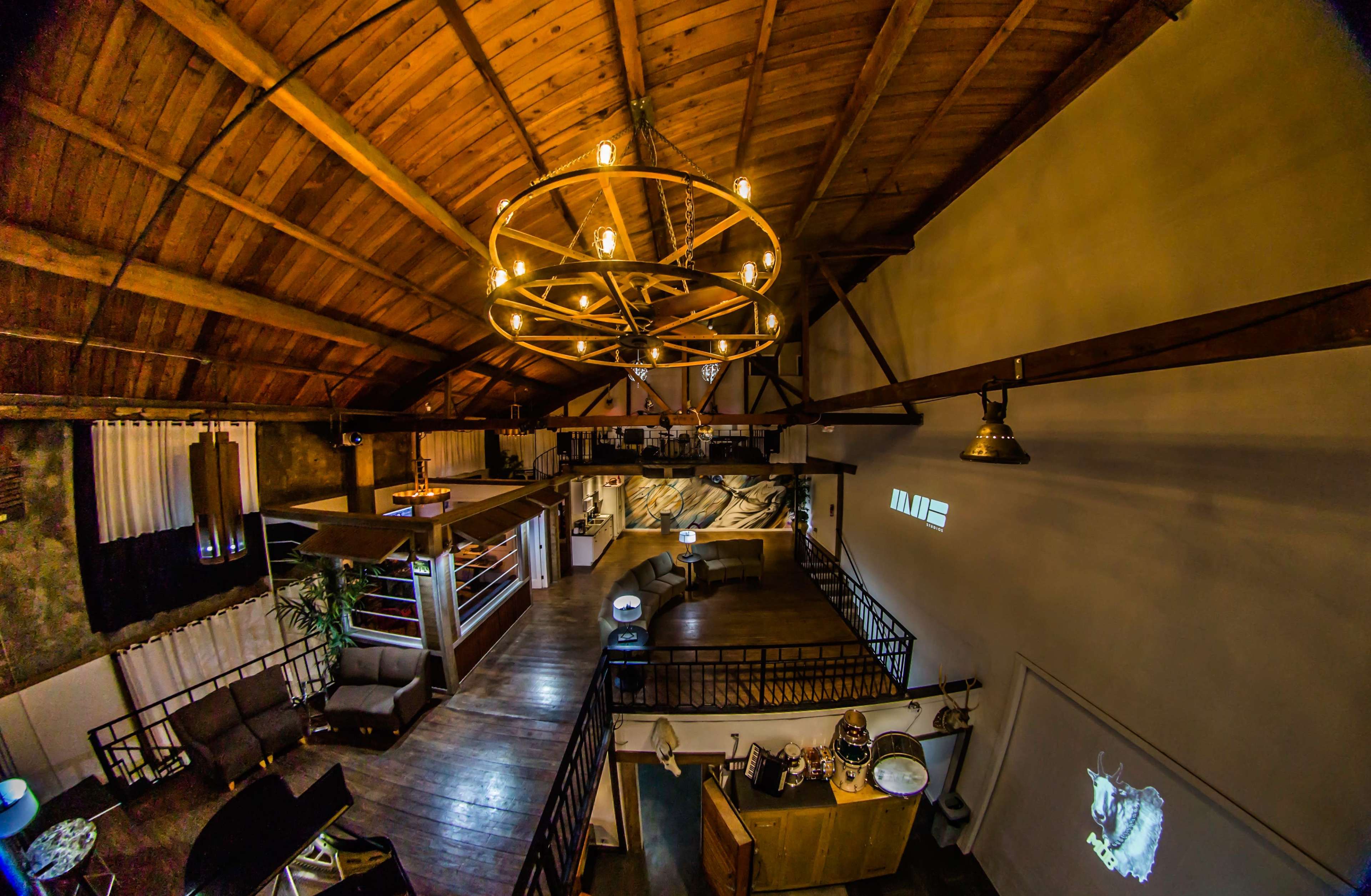 The interior of a spacious loft features exposed wooden beams, a large circular chandelier, and various seating areas arranged around a central open space.