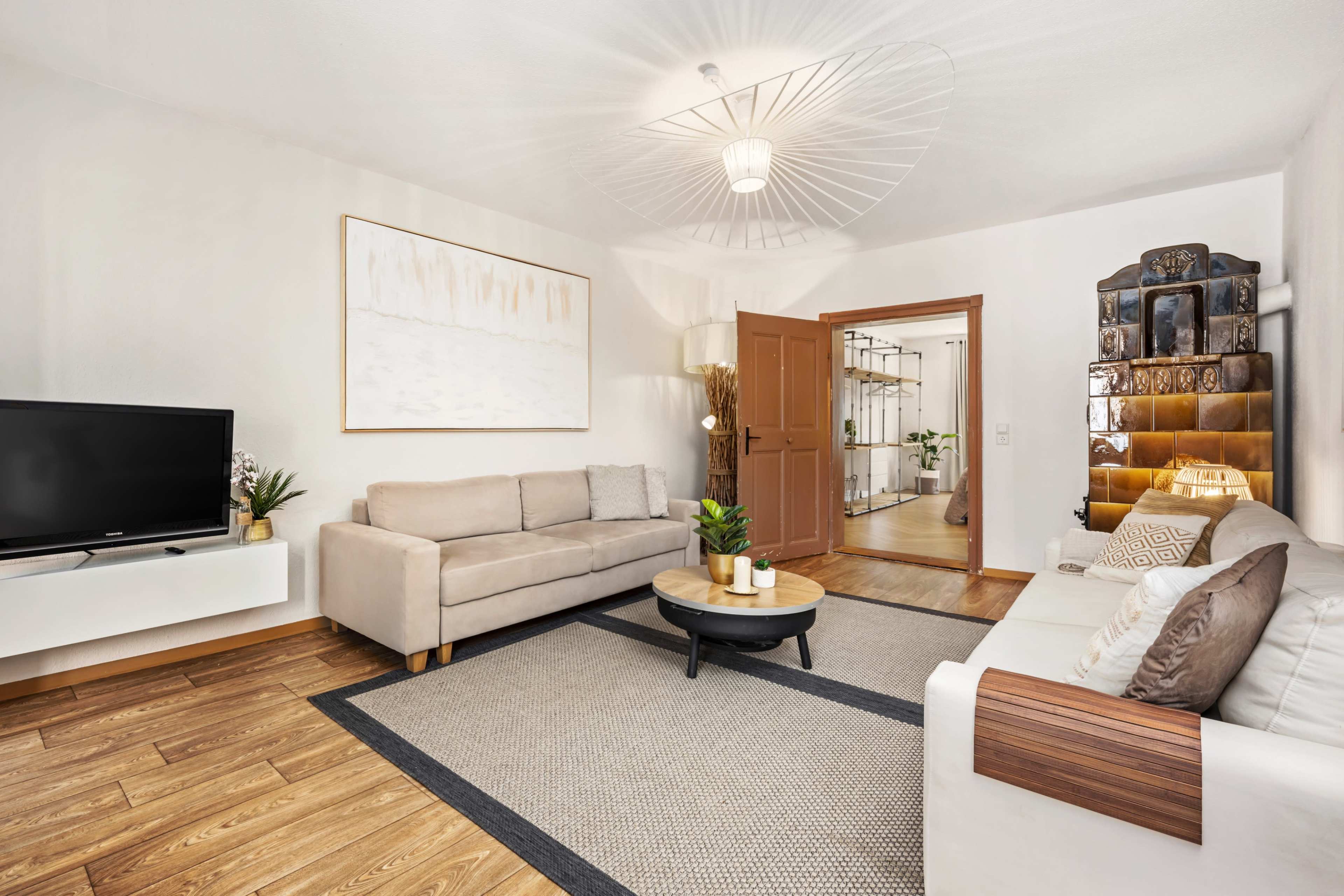 A modern living room features a beige sofa, a white couch, a round coffee table, and a television, with an open doorway leading to a bright space.