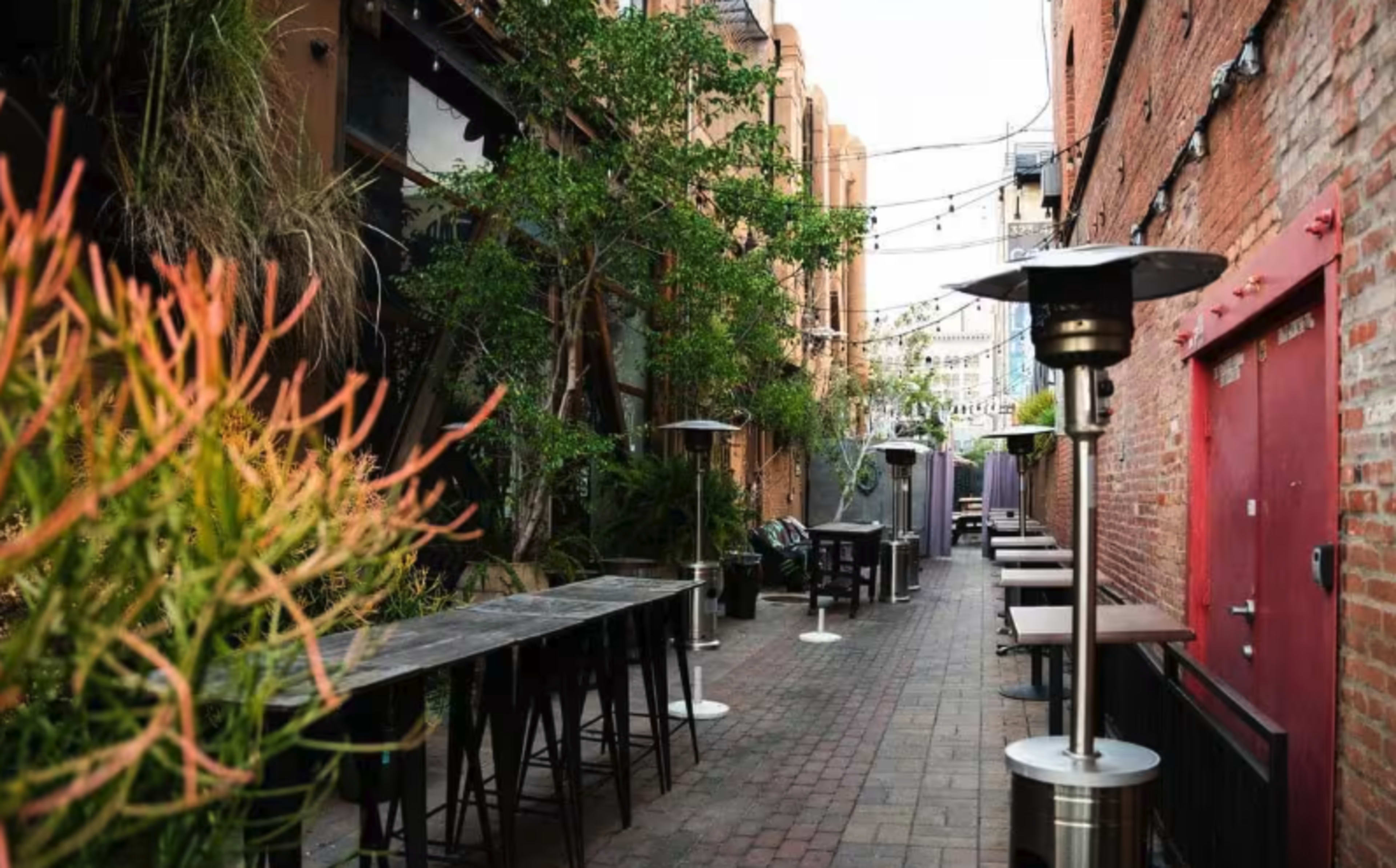 The image shows a narrow alleyway with tables and heat lamps on either side, flanked by greenery and brick walls.