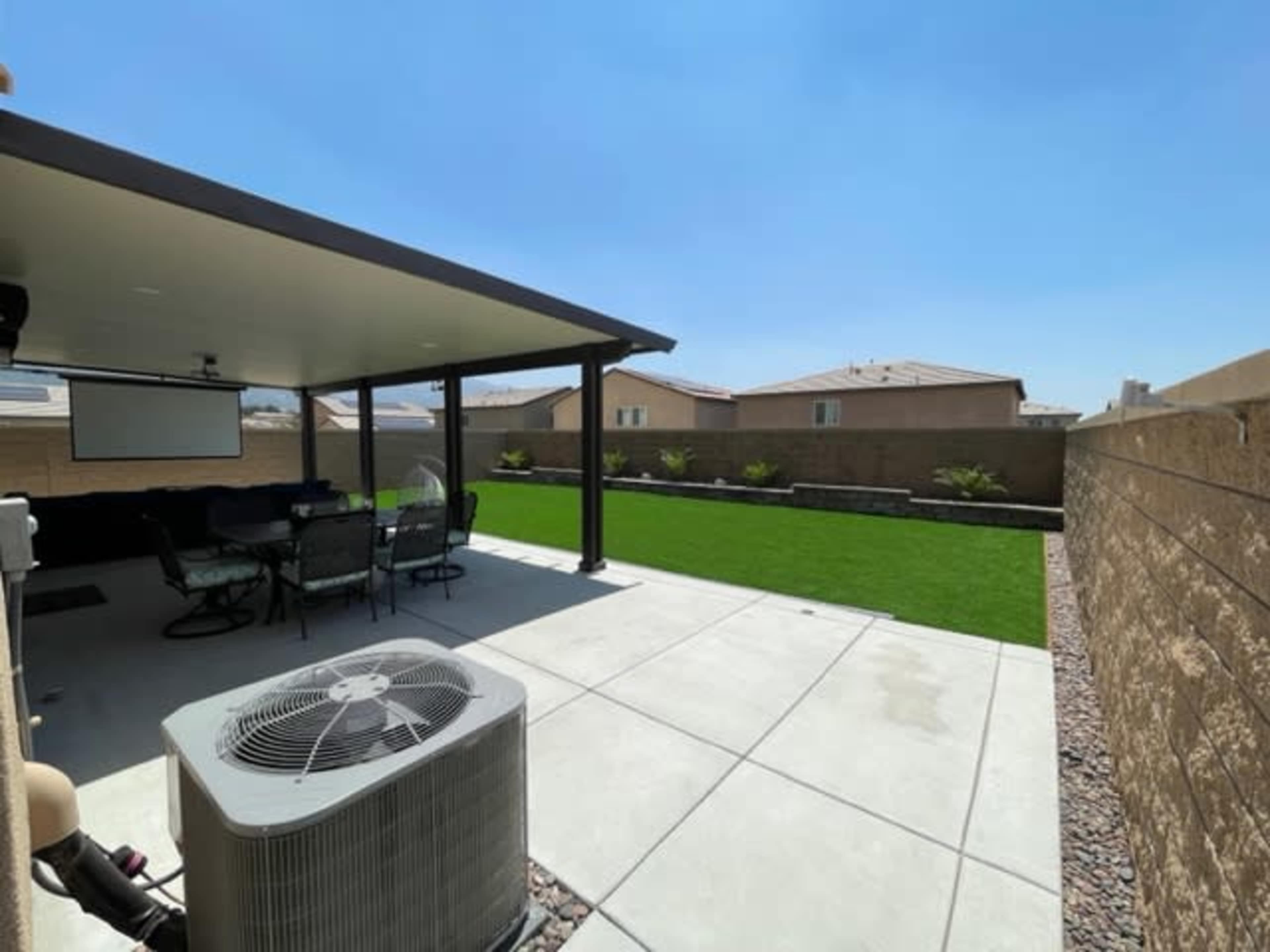 The image shows a back patio area with a covered seating space, an air conditioning unit, and a grassy lawn enclosed by a fence.