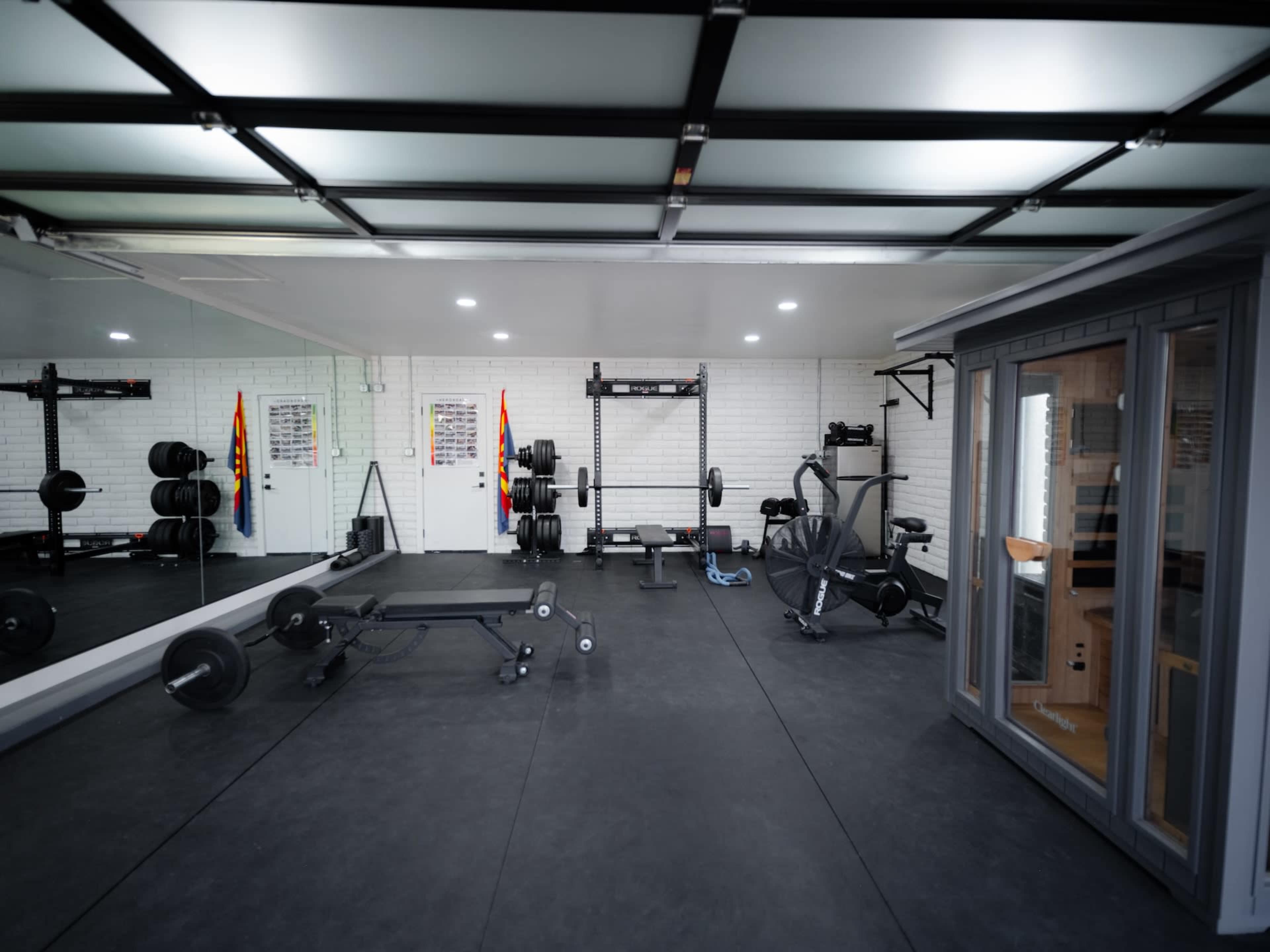 The image shows a spacious gym with various workout equipment, including free weights, a bench press, and a sauna, all set on a black rubber floor.