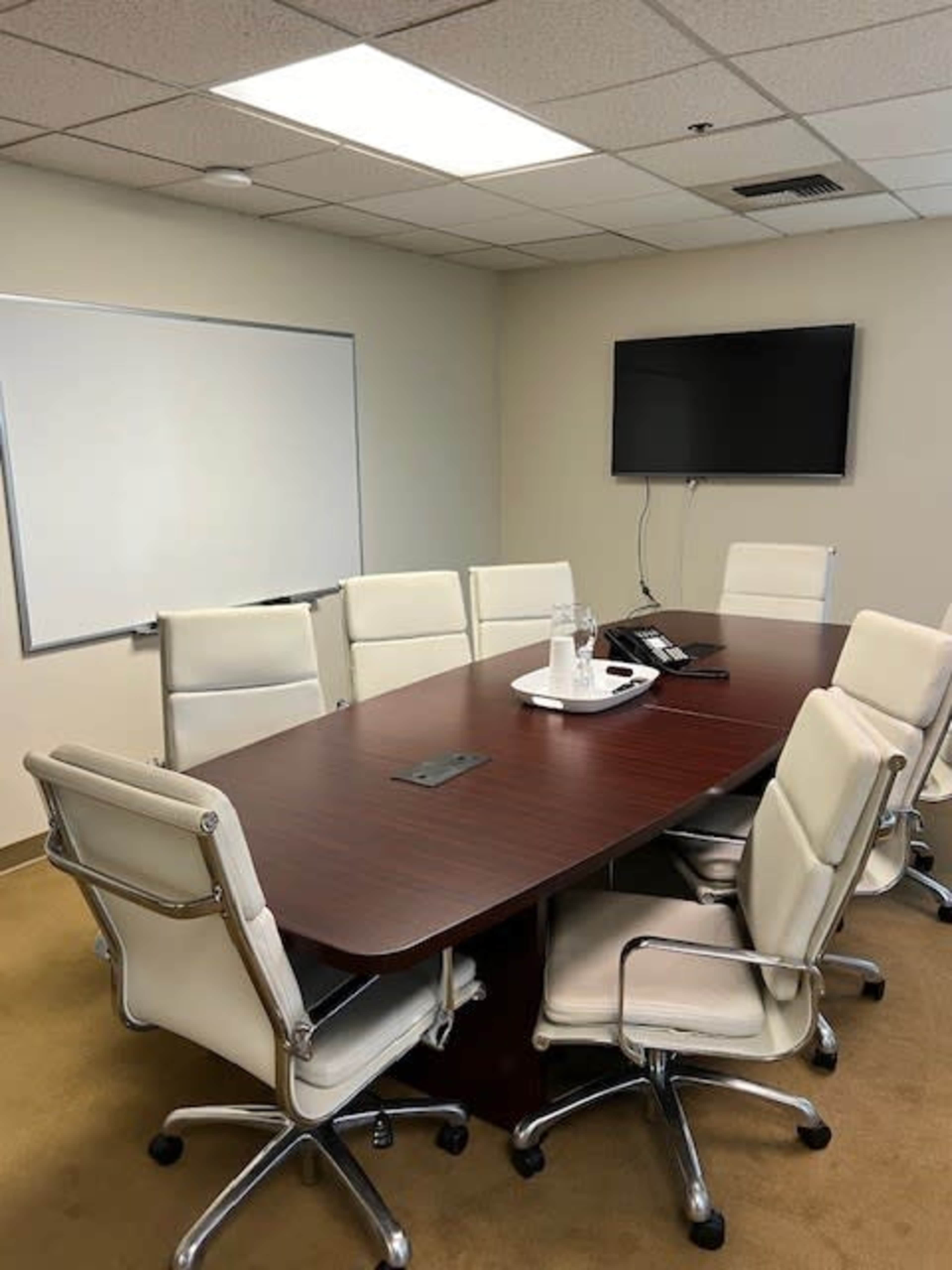 The image shows a conference room with a large wooden table surrounded by eight white chairs, a flat-screen TV mounted on the wall, and a whiteboard.