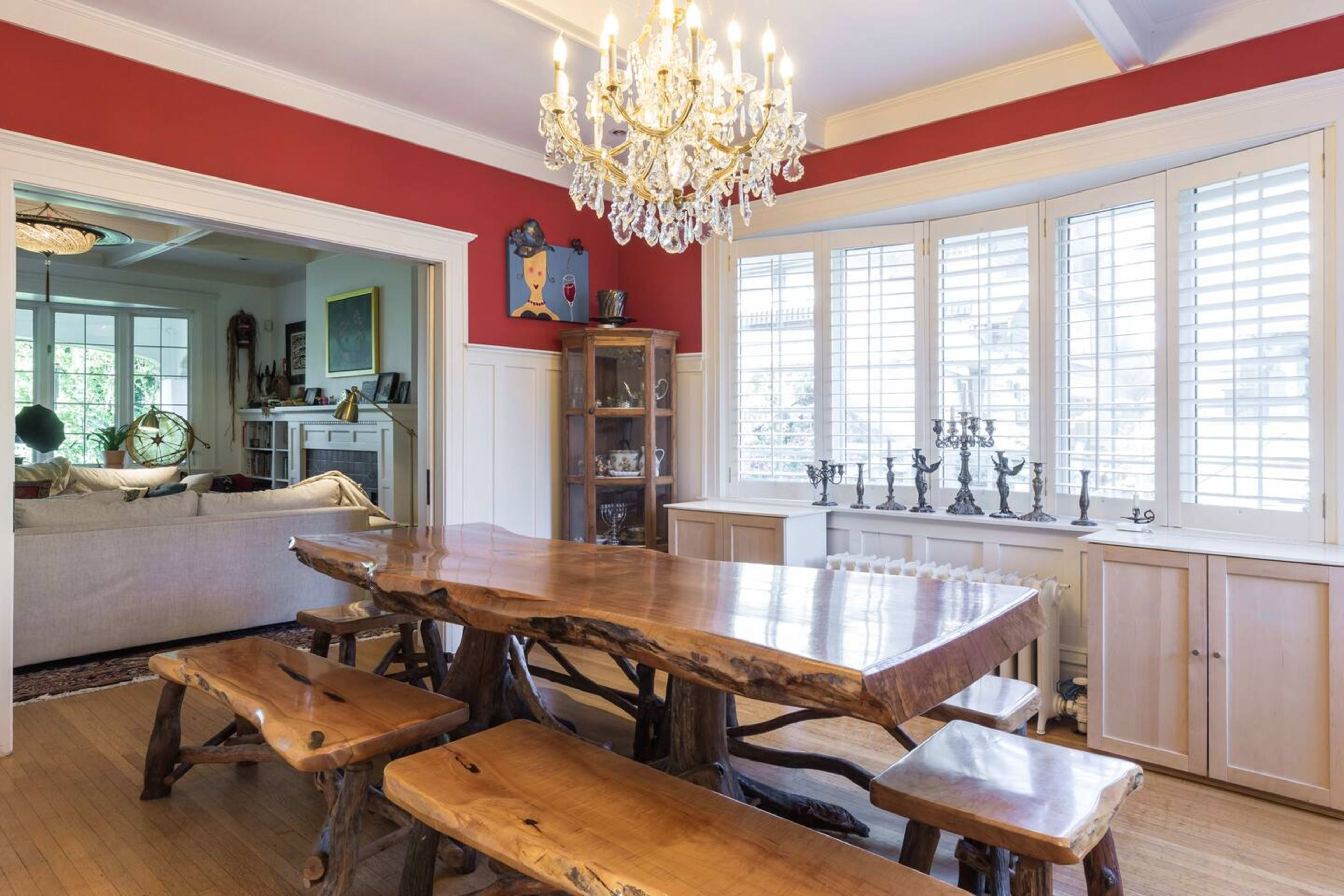 A large wooden dining table with matching benches is set in a red-walled dining area featuring a chandelier and large windows with shutters.