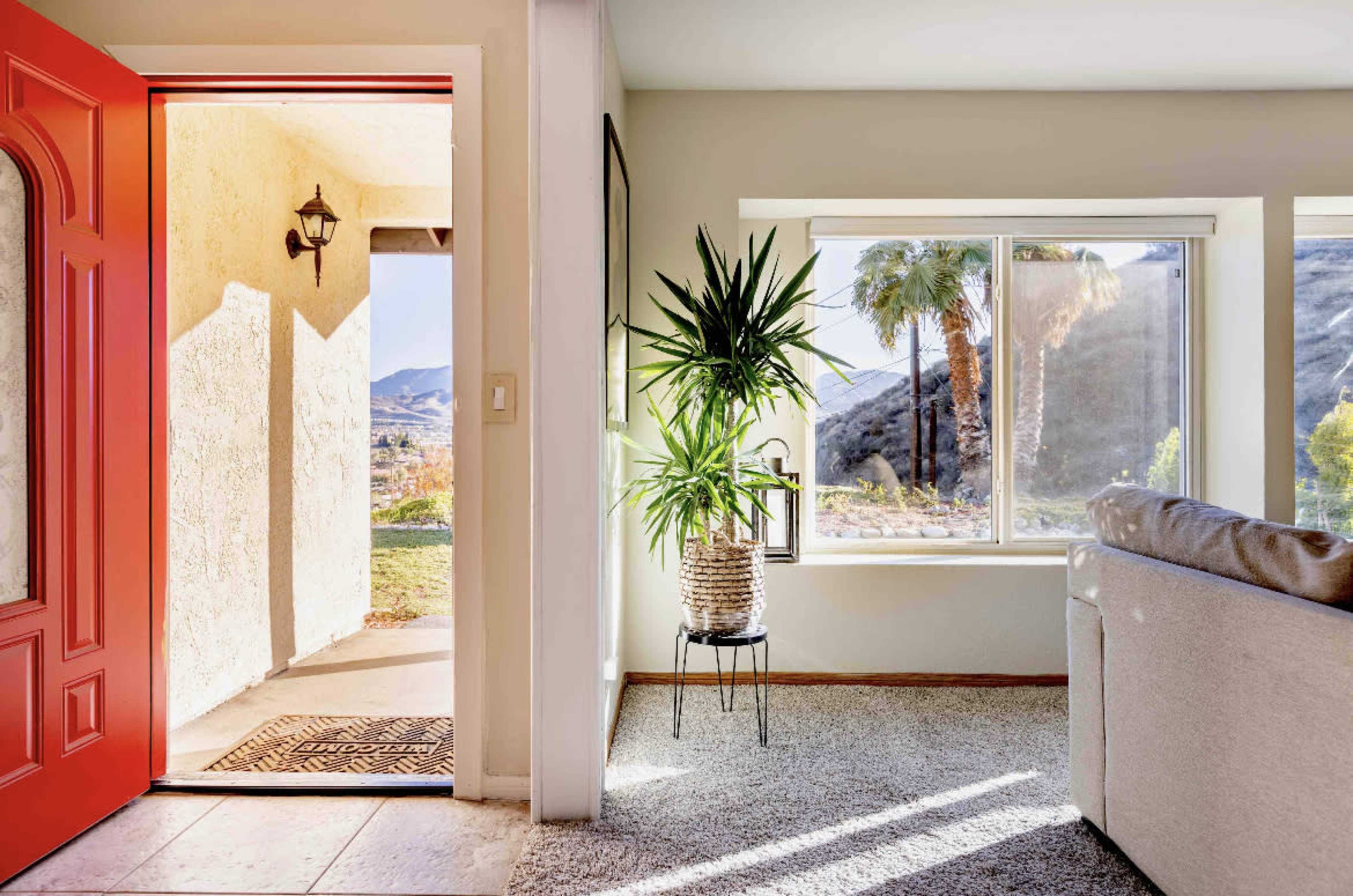 A brightly lit entryway features a vibrant red front door, leading into a cozy living room with a potted plant by the window.