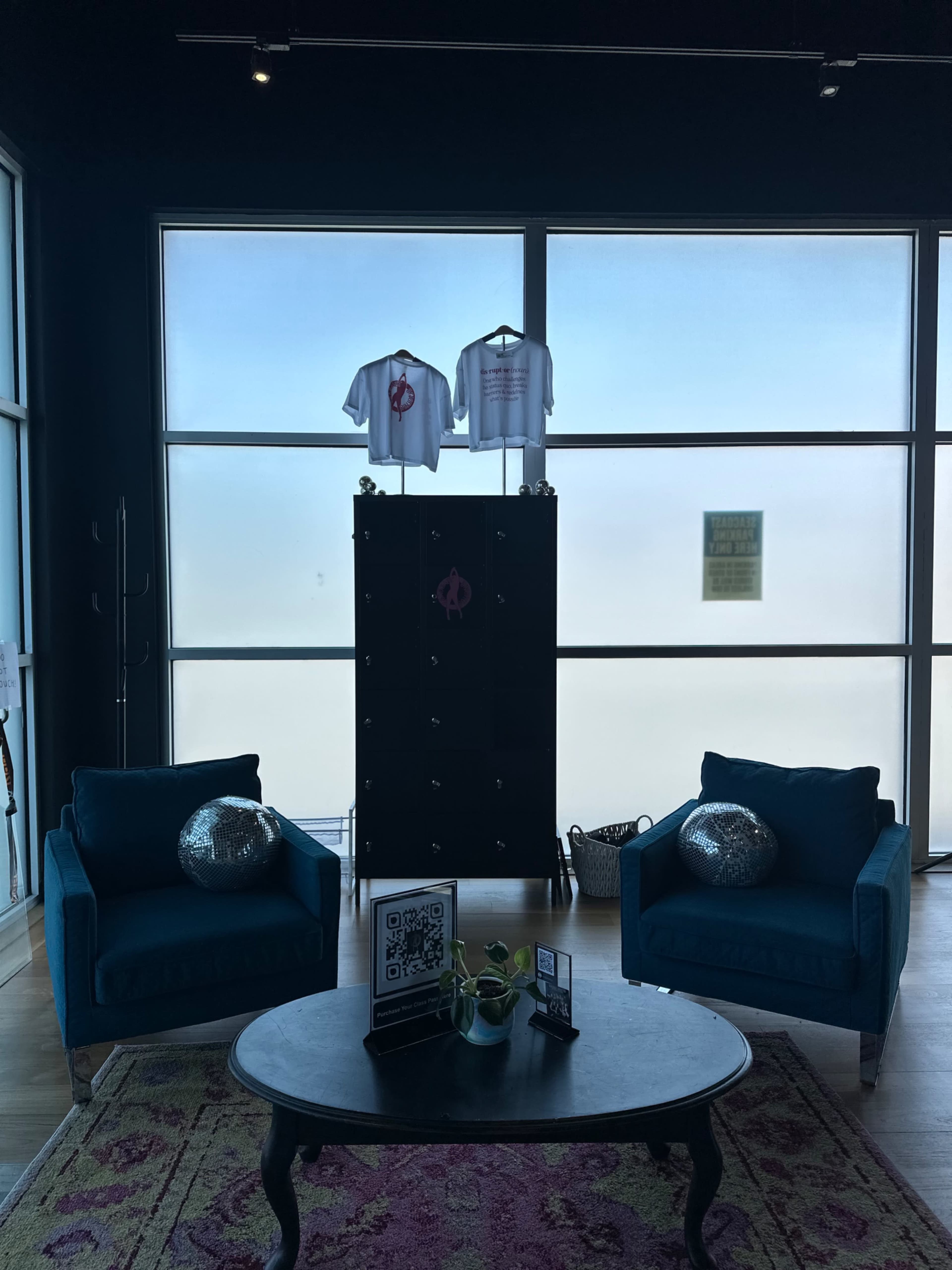 The image shows a modern indoor space featuring two blue chairs facing a round table, with a large window behind displaying a clear view of the sky, and clothing displayed on hangers nearby.