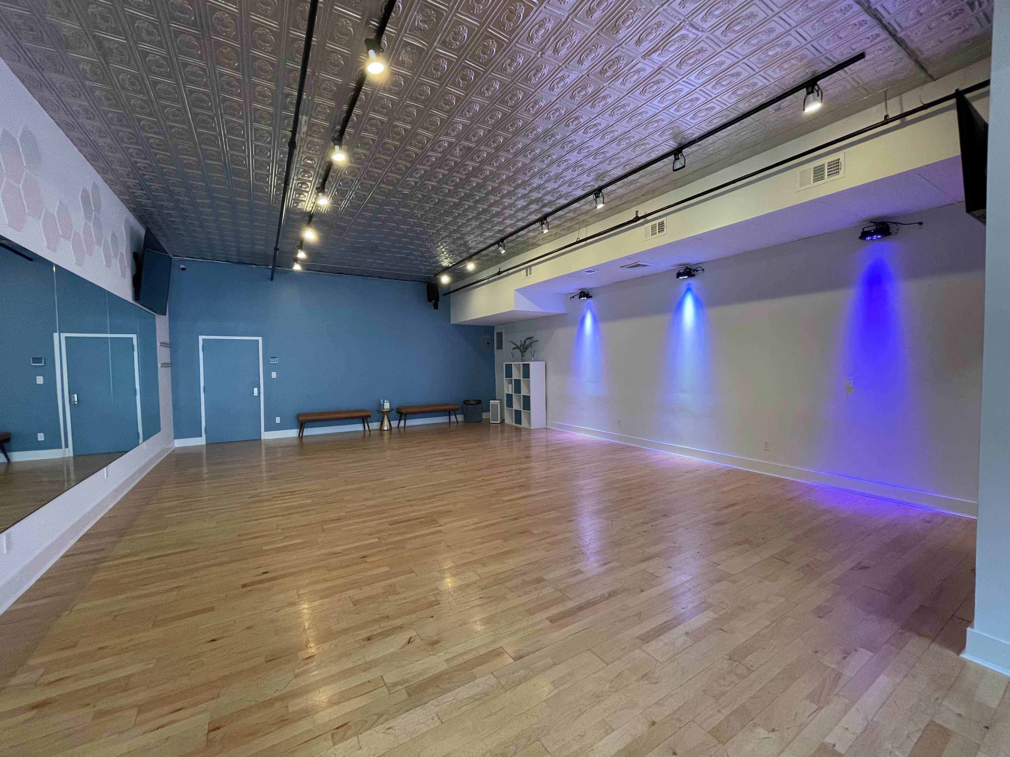 The image shows a spacious, uncluttered dance studio with wooden flooring, a large mirror, and blue accent lighting.