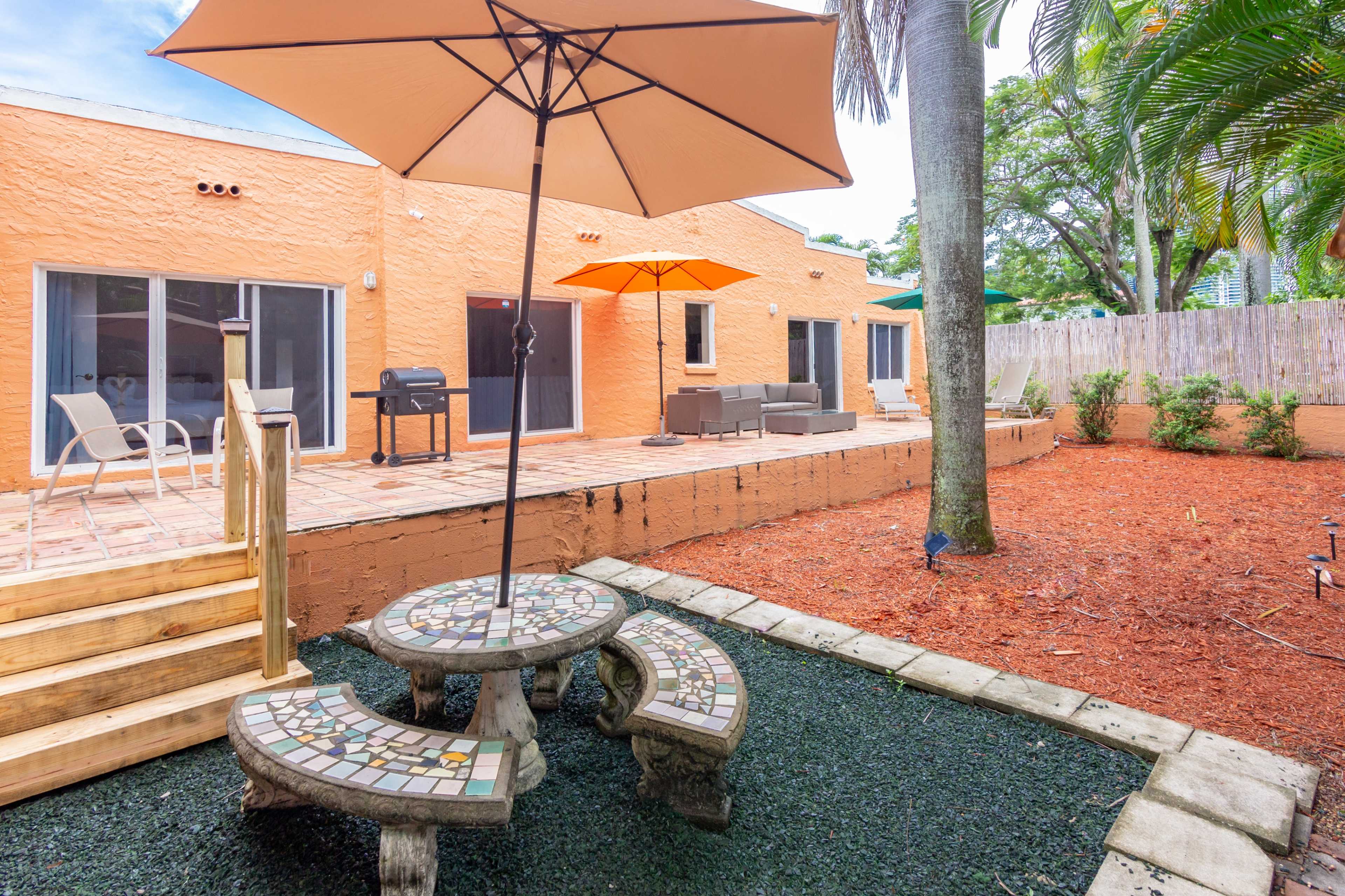 A backyard patio area with a mosaic table and seating, surrounded by mulch and grass, with a grill and lounge furniture against a colorful wall.
