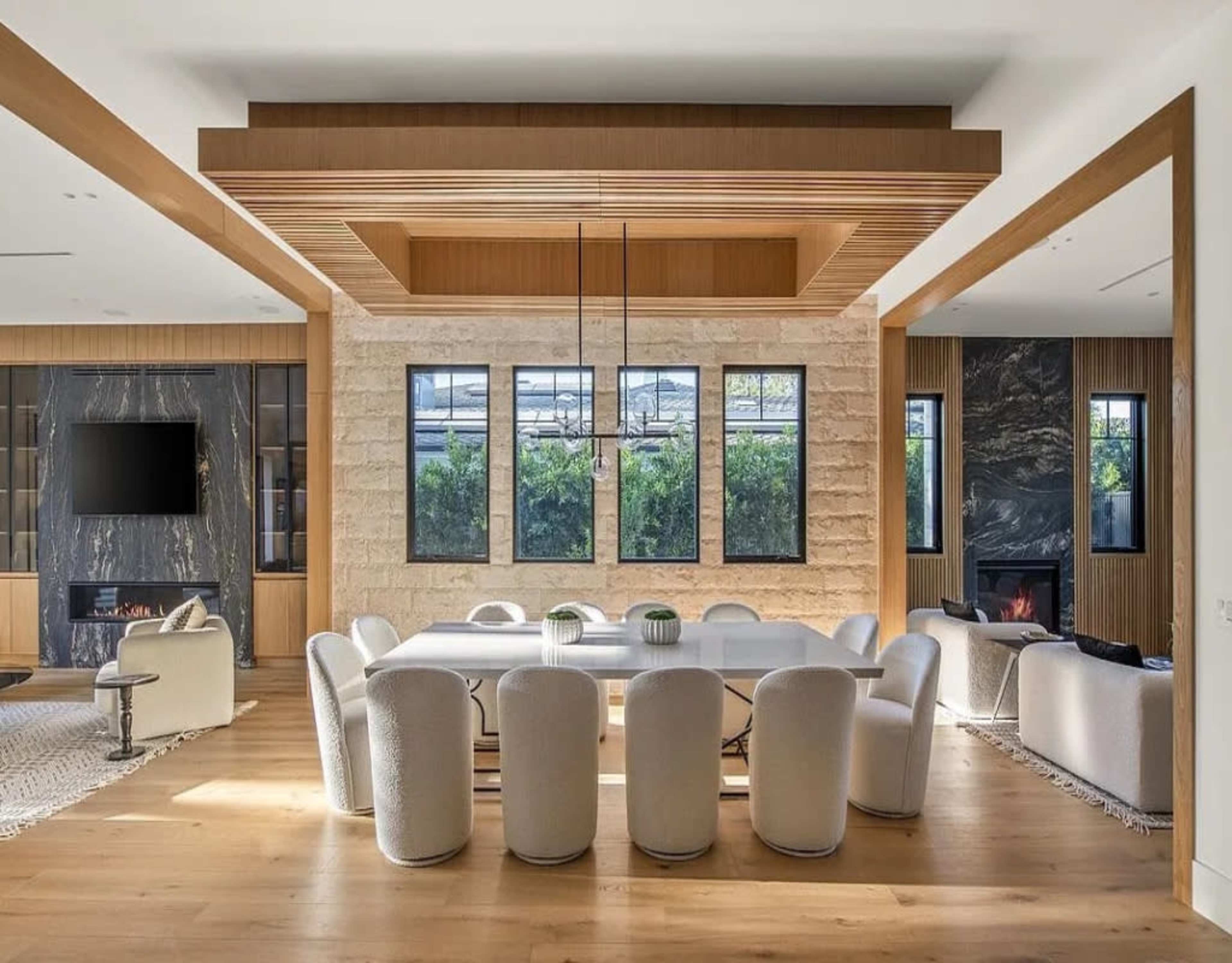 A spacious dining area features a large table surrounded by upholstered chairs, with a textured stone wall and large windows offering views of greenery.