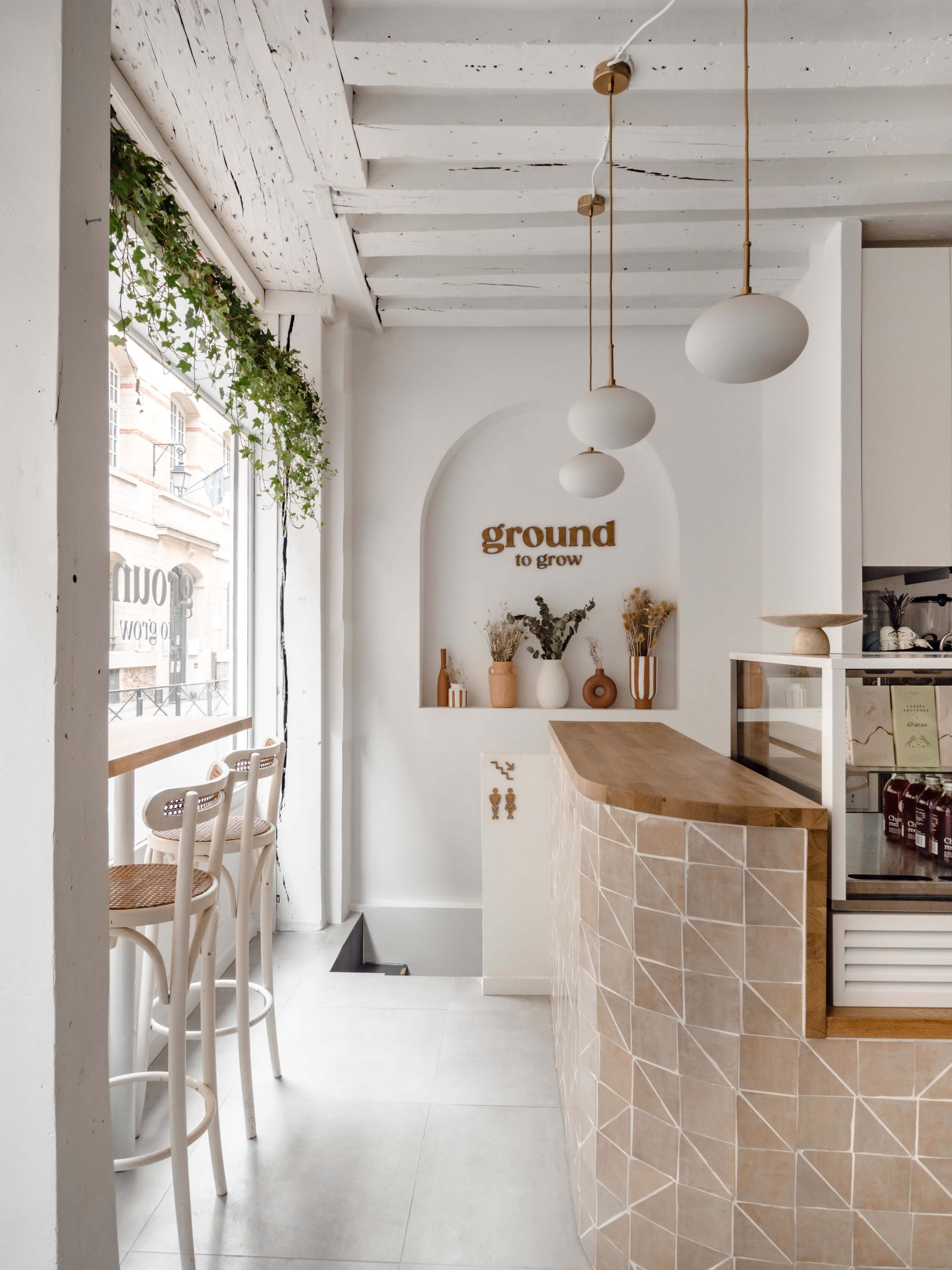 The image shows a bright café interior with a light wooden counter, white shelves, and a plant hanging from the ceiling.