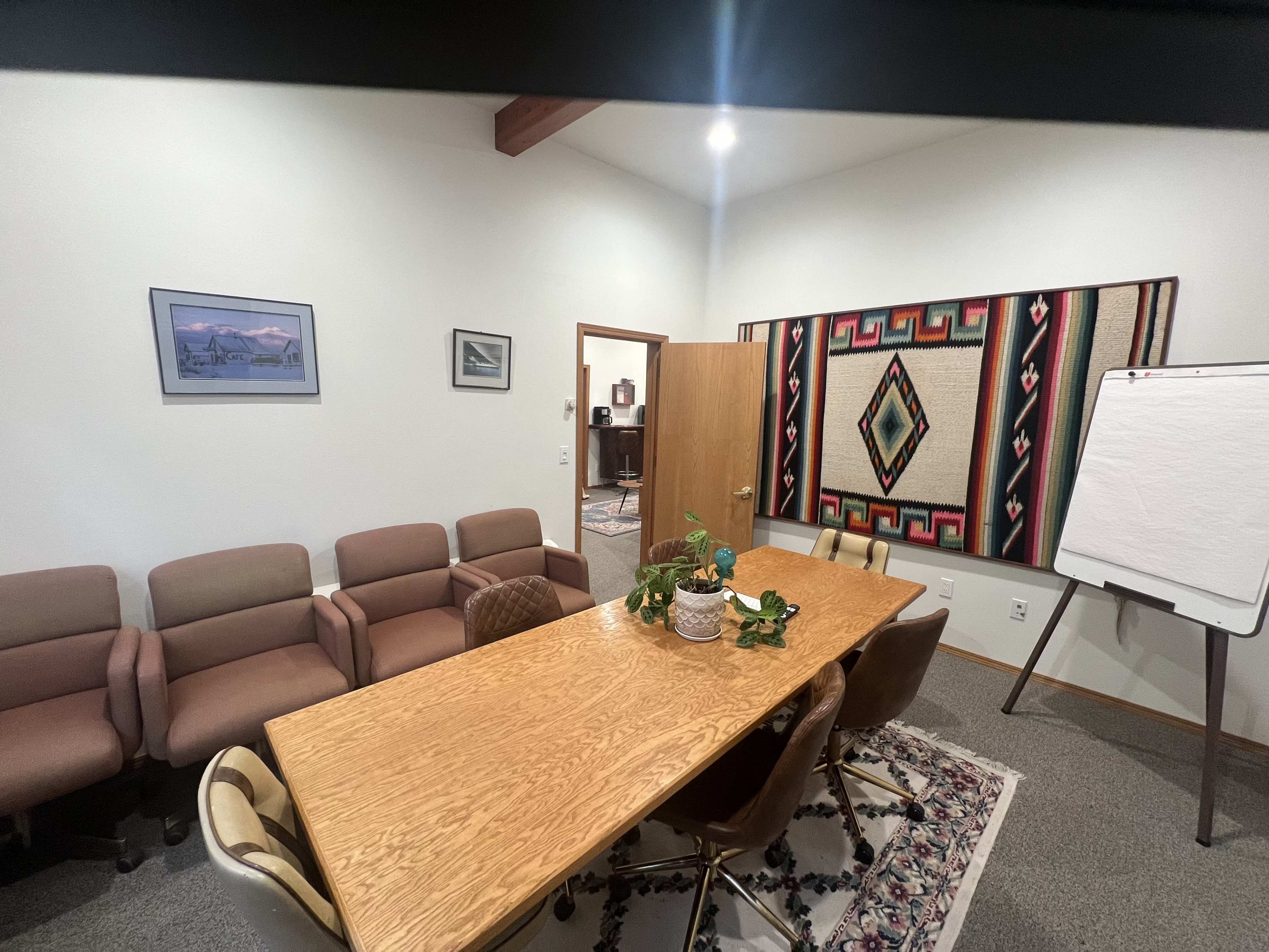 A conference room features a wooden table surrounded by chairs, with a wall decorated by a large textile and two framed pictures.