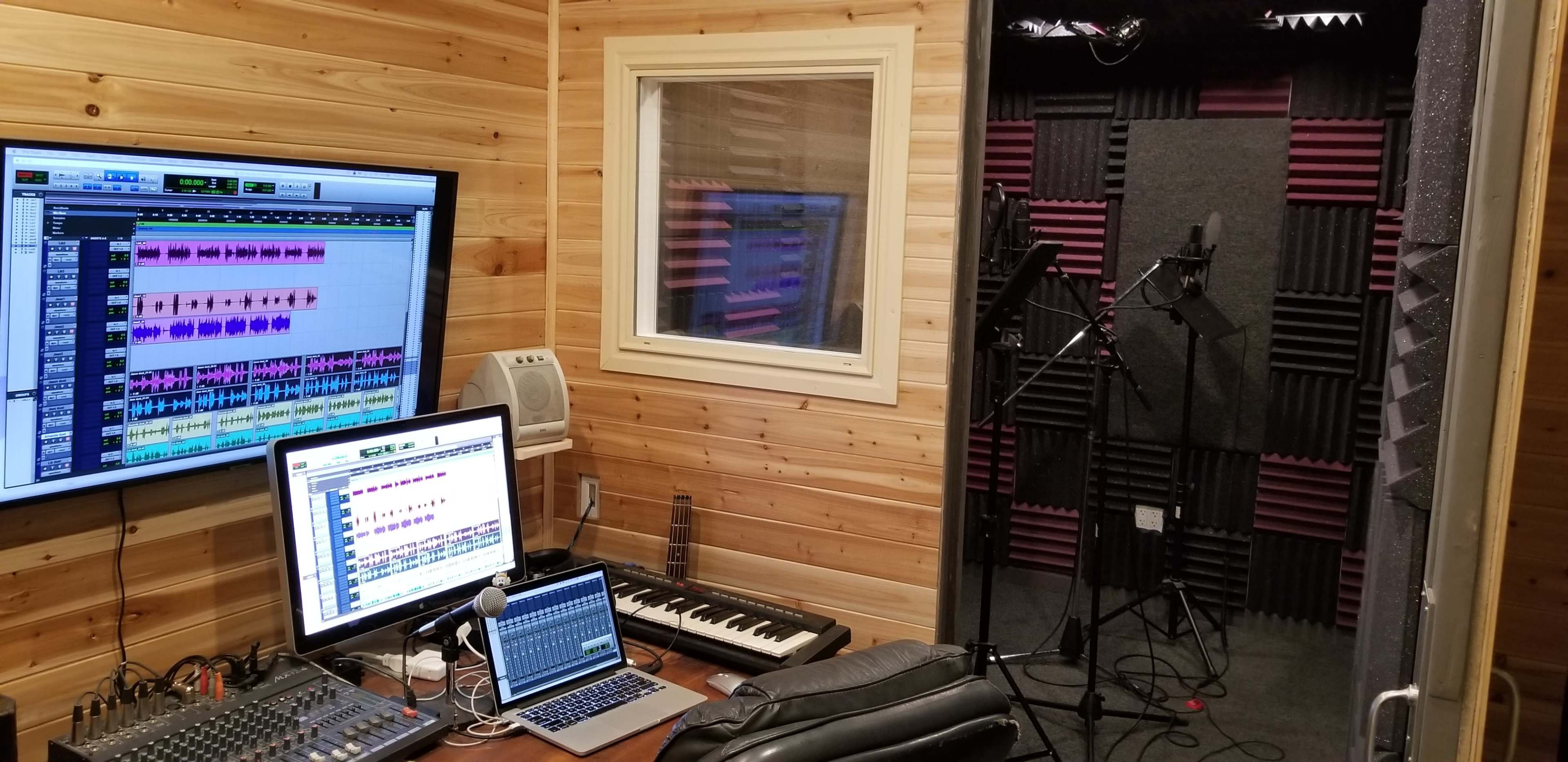 A sound recording studio with a computer setup, a keyboard, and a vocal booth in the background.