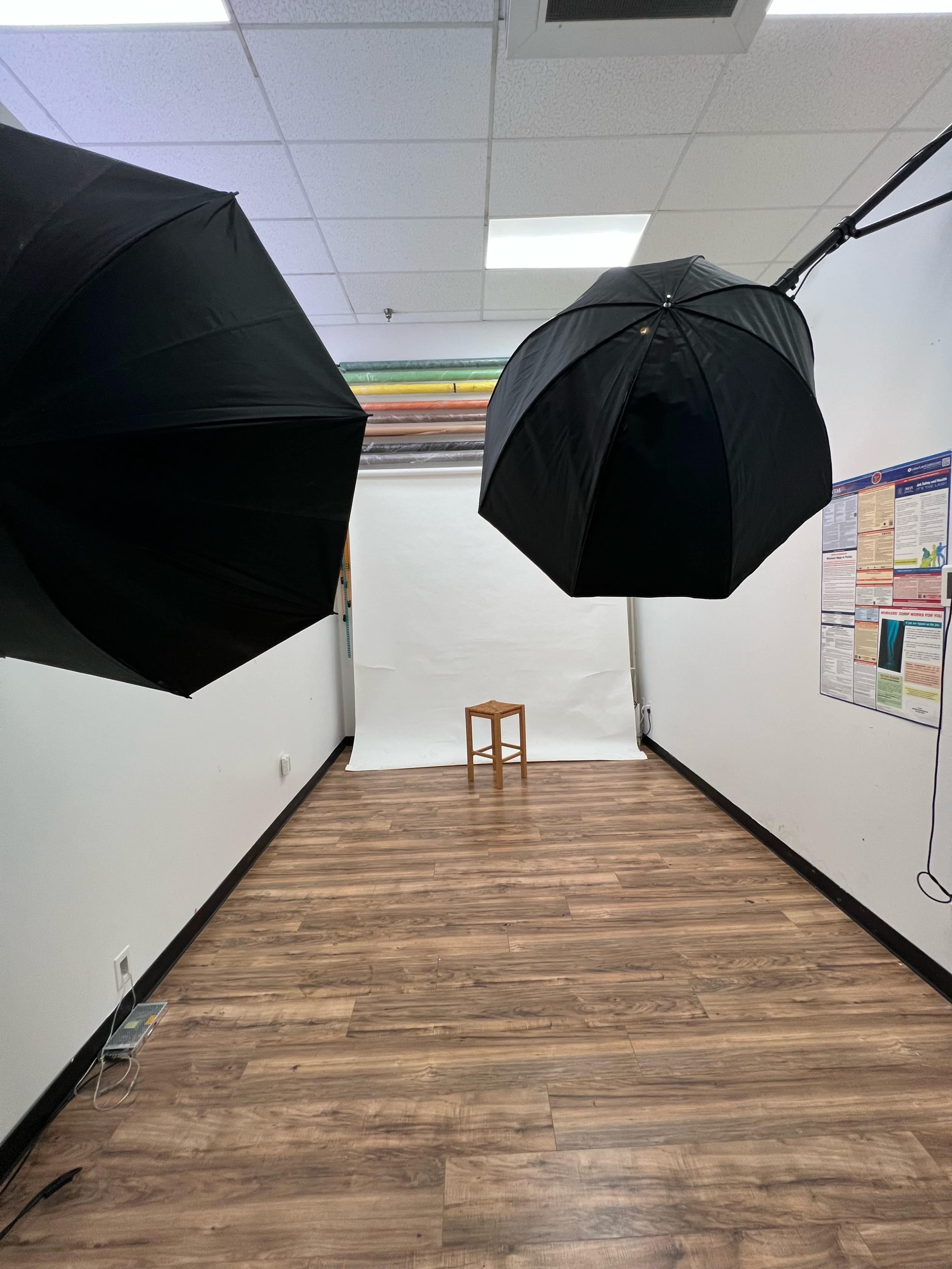 The photo shows an empty studio space with two large black umbrellas positioned overhead and a wooden stool in the center, facing a white backdrop.