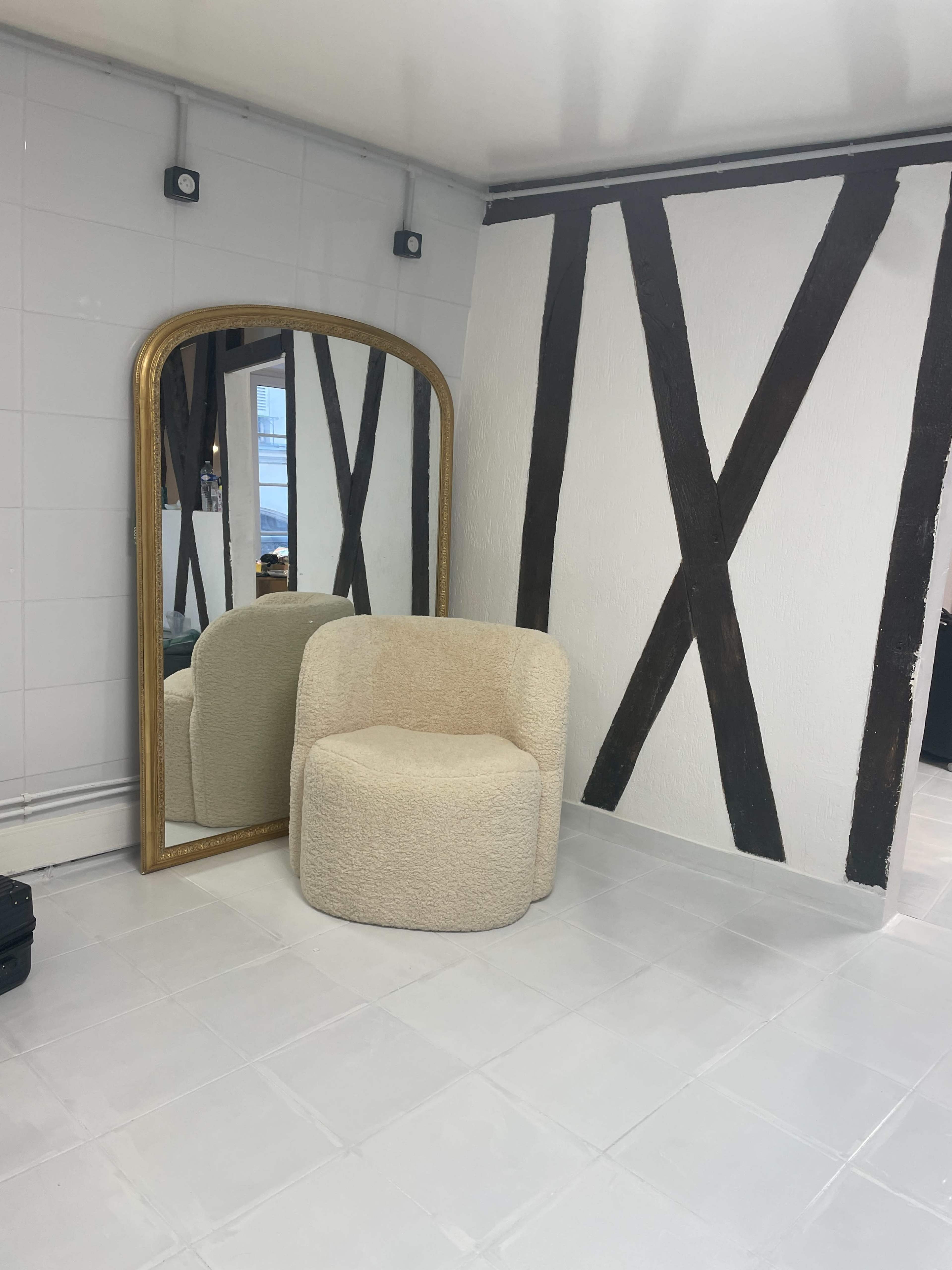 The image shows a room with a large arched mirror, a short, round chair upholstered in light fabric, and a wall featuring dark wooden beams.